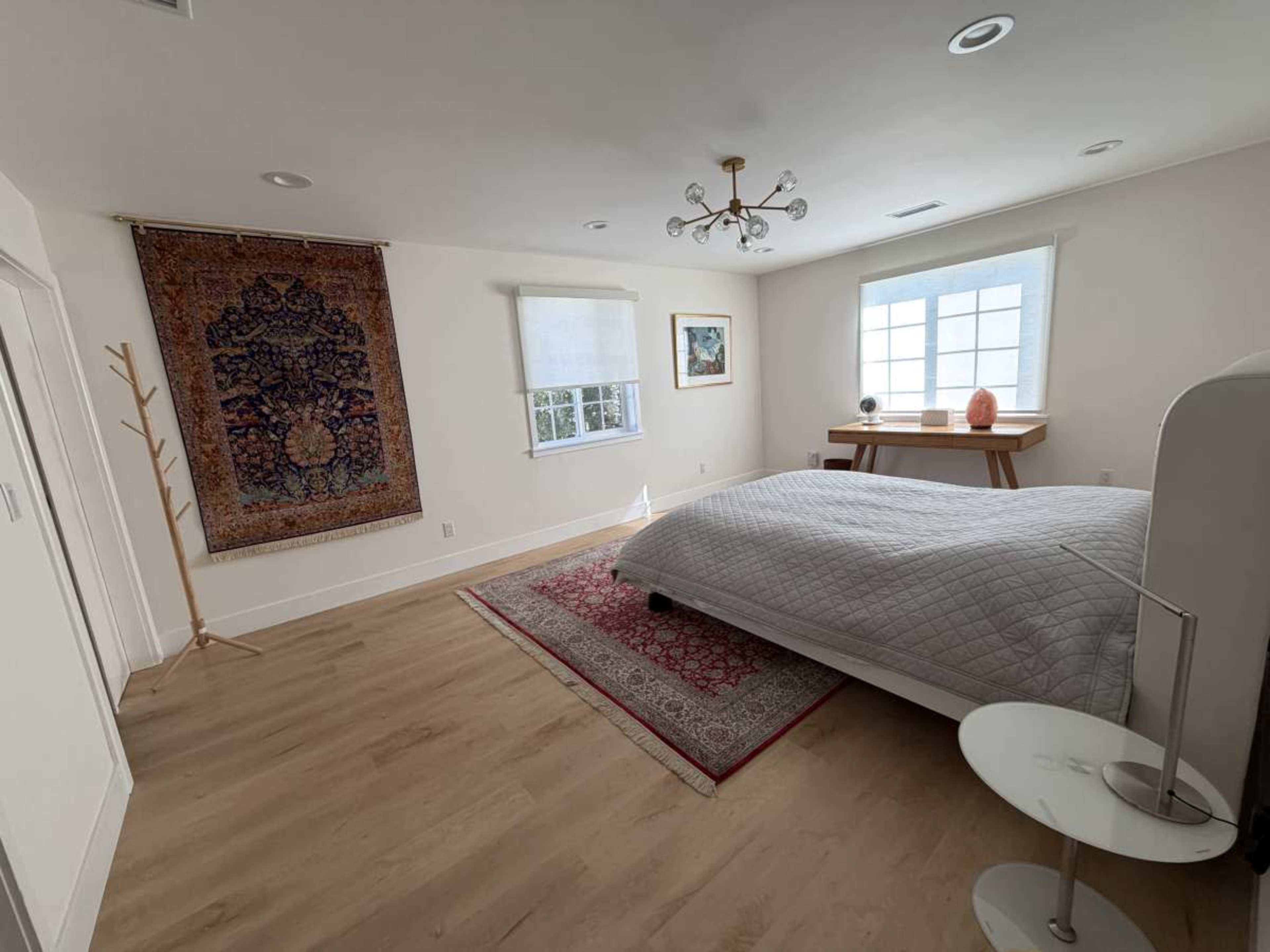 The image shows a bright, minimalist bedroom featuring a bed, a decorative rug, a wall tapestry, a small side table, and a large window with natural light.
