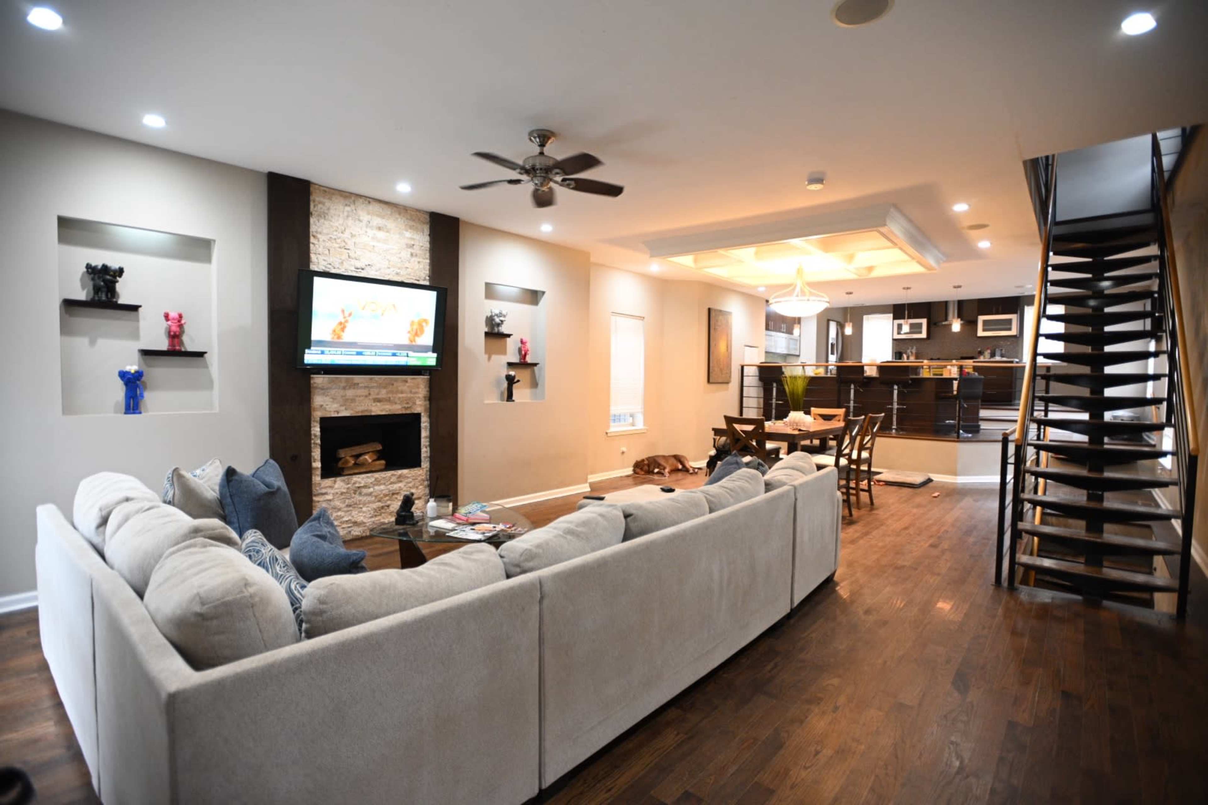 A modern living room with a beige sectional couch, a stone fireplace with a TV above it, and a staircase leading to an upper level.