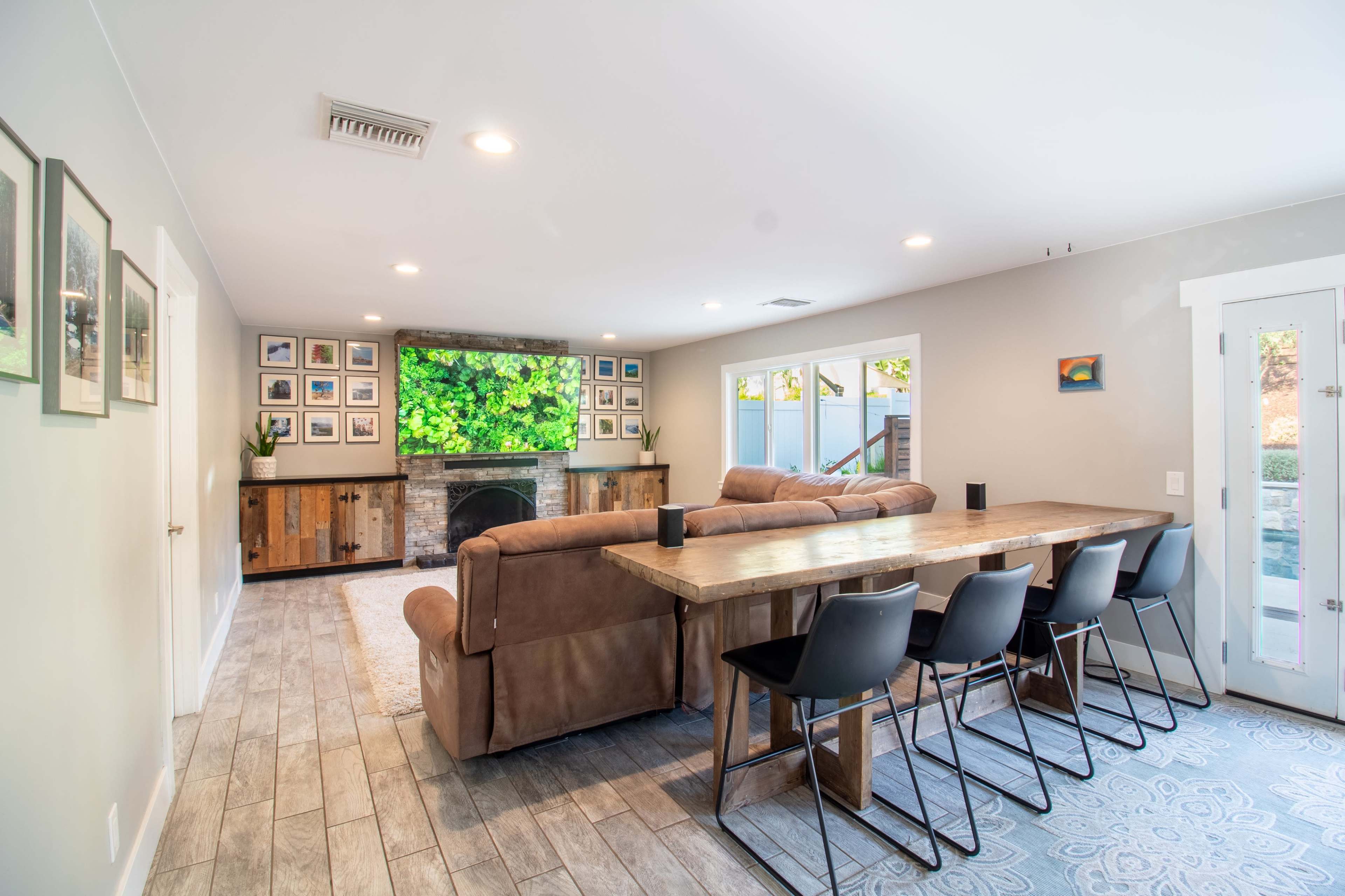 A living space with a large sectional sofa facing a mounted television above a stone fireplace, complemented by a long dining table with black chairs and framed photos on the walls.