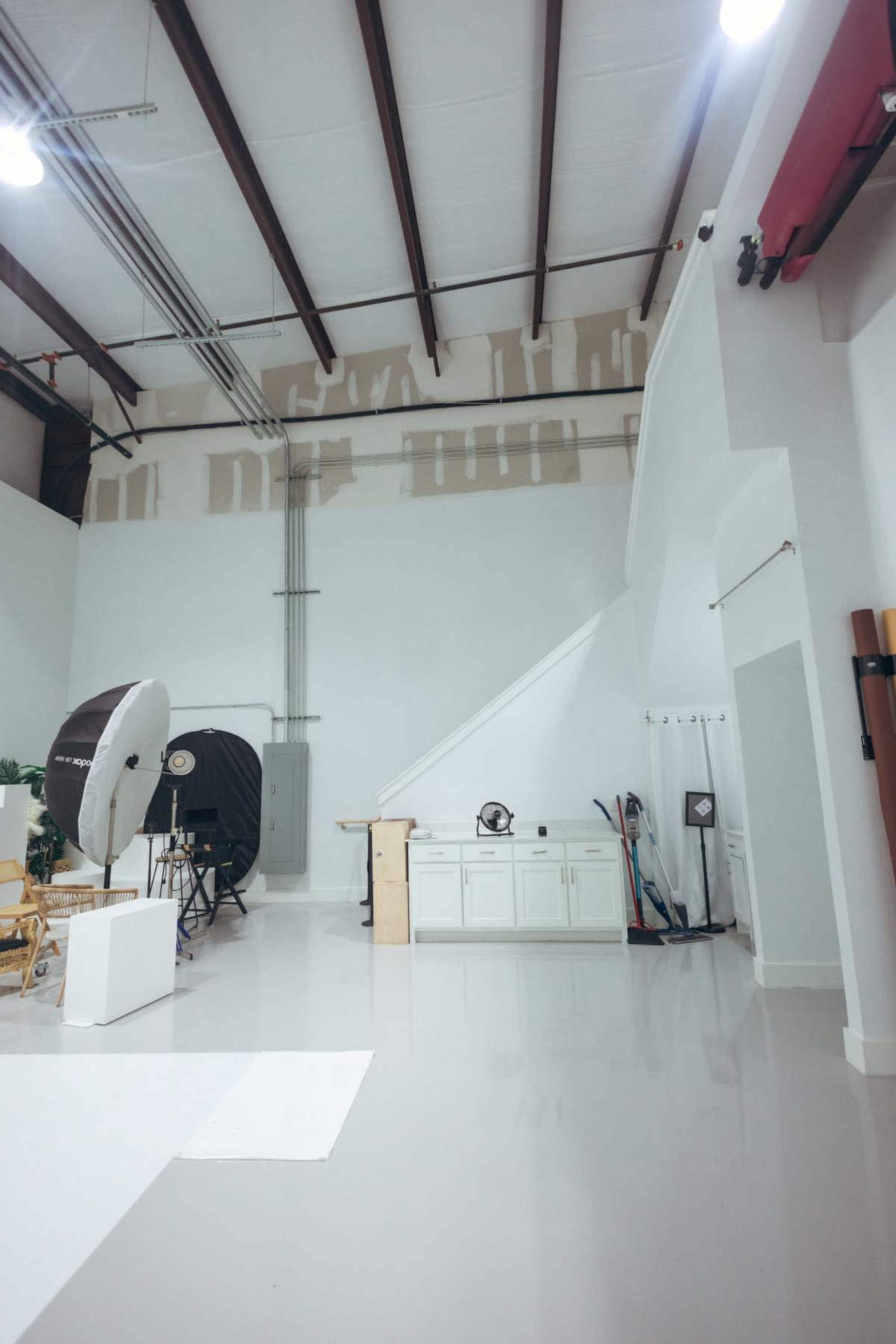 The image shows an interior space with a high ceiling, featuring painted walls, industrial light fixtures, and various photography equipment arranged around the room.