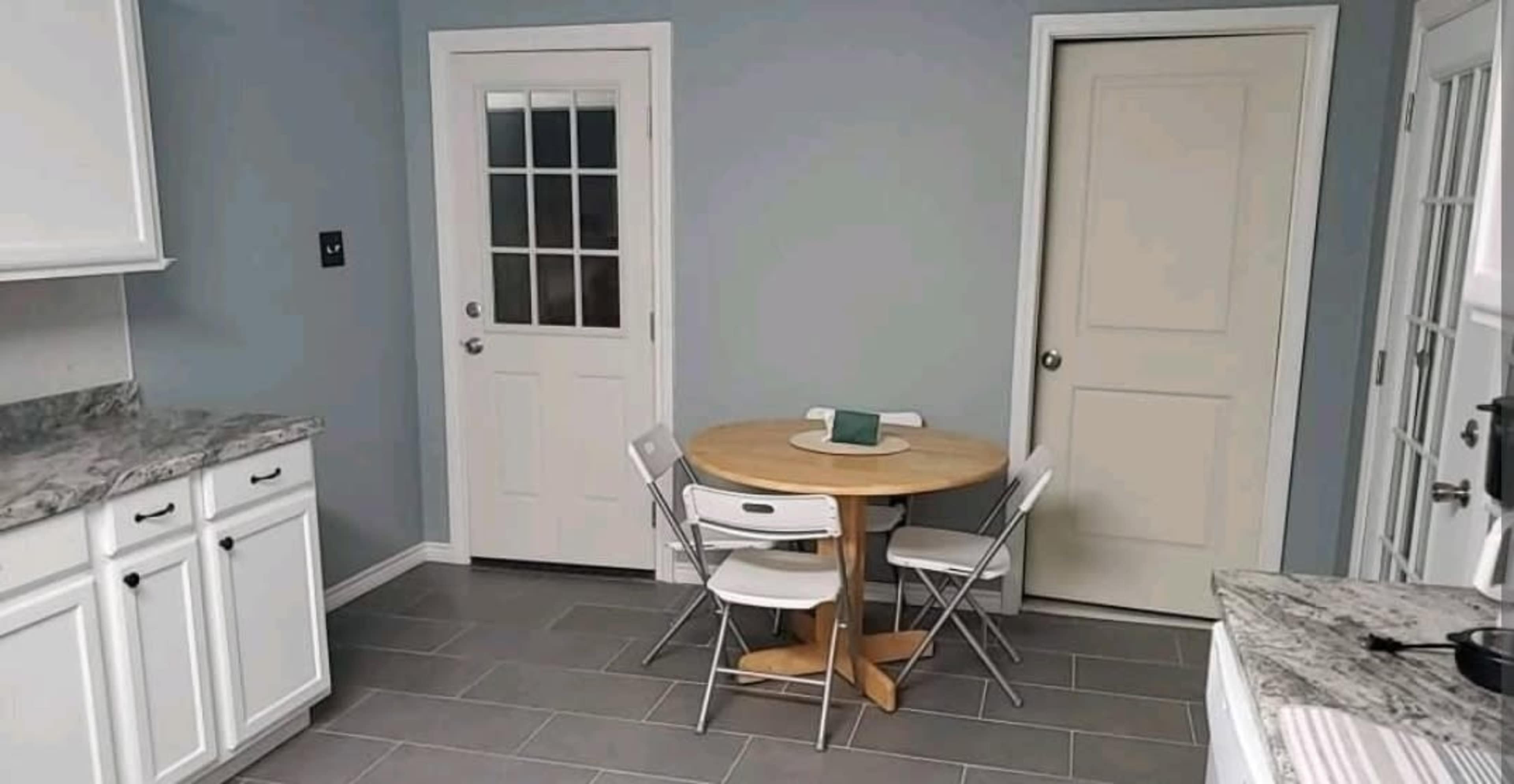 A small kitchen features a round wooden table with four chairs, a door leading outside, and a door leading to another room, all set against light blue walls and gray tiled flooring.