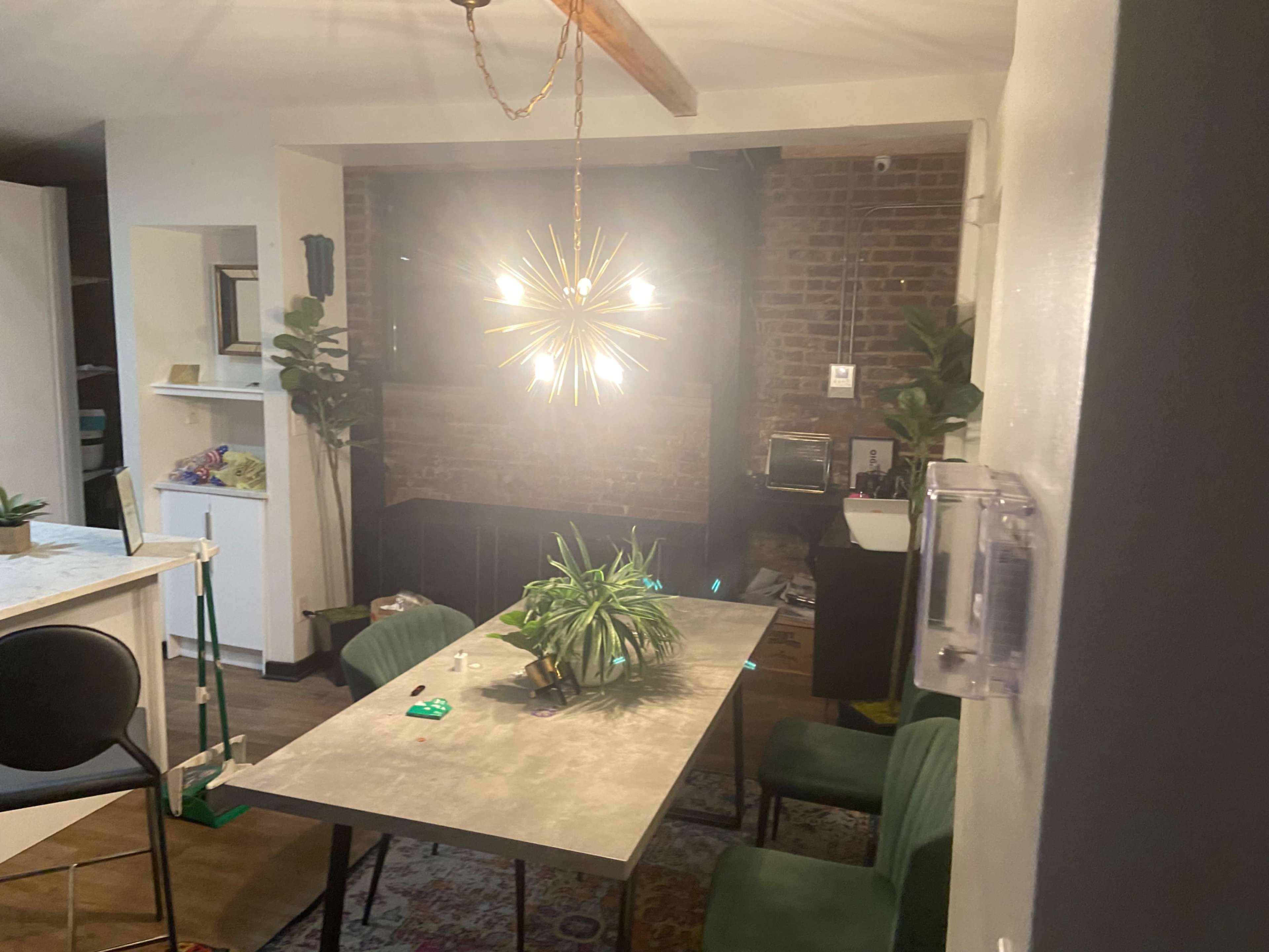 A dining area with a modern light fixture hanging above a table, surrounded by plants and furniture against a brick wall.