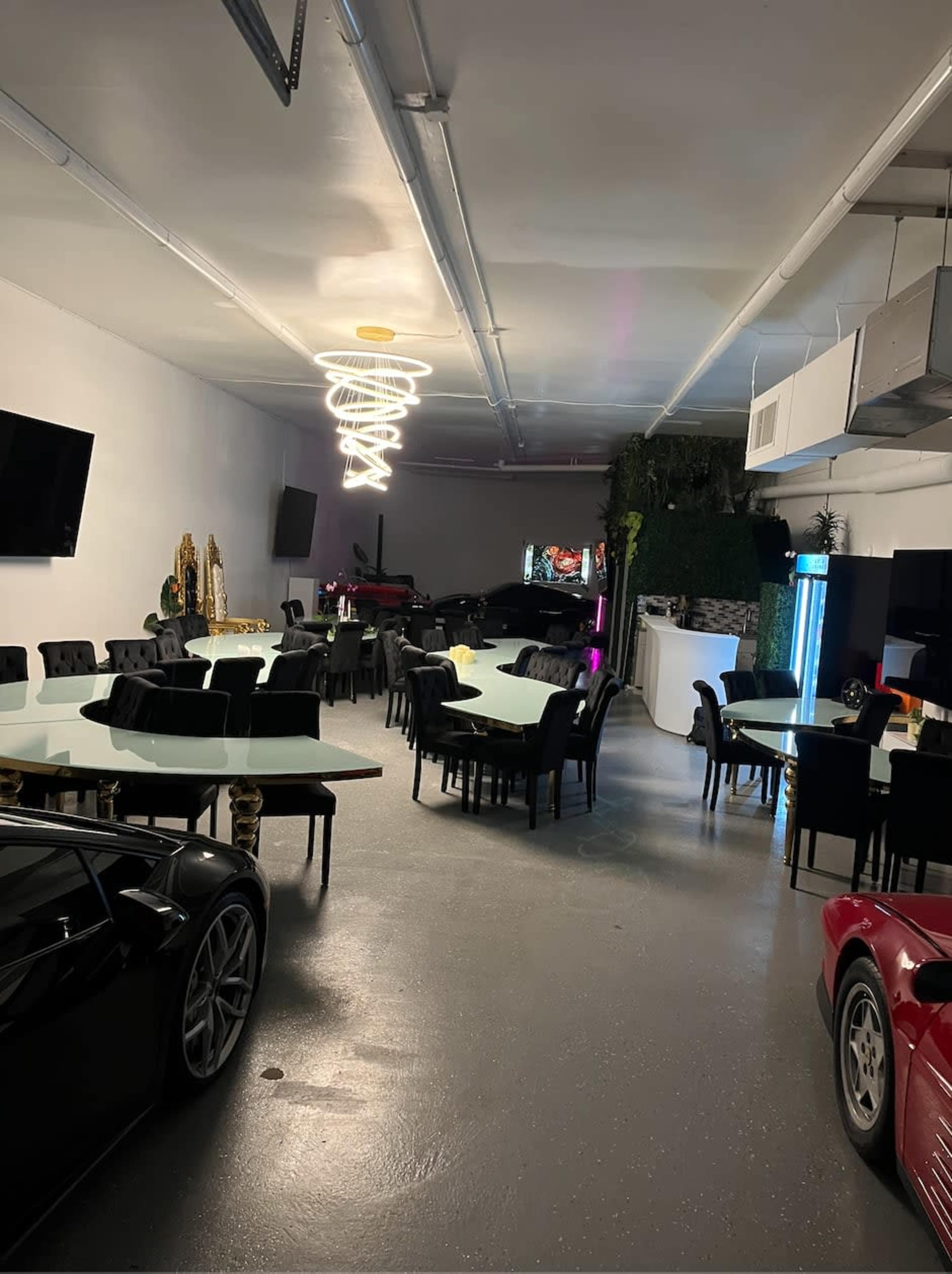 The image shows a spacious garage that has been converted into a dining area, featuring multiple tables and chairs, along with luxury cars parked along the sides.