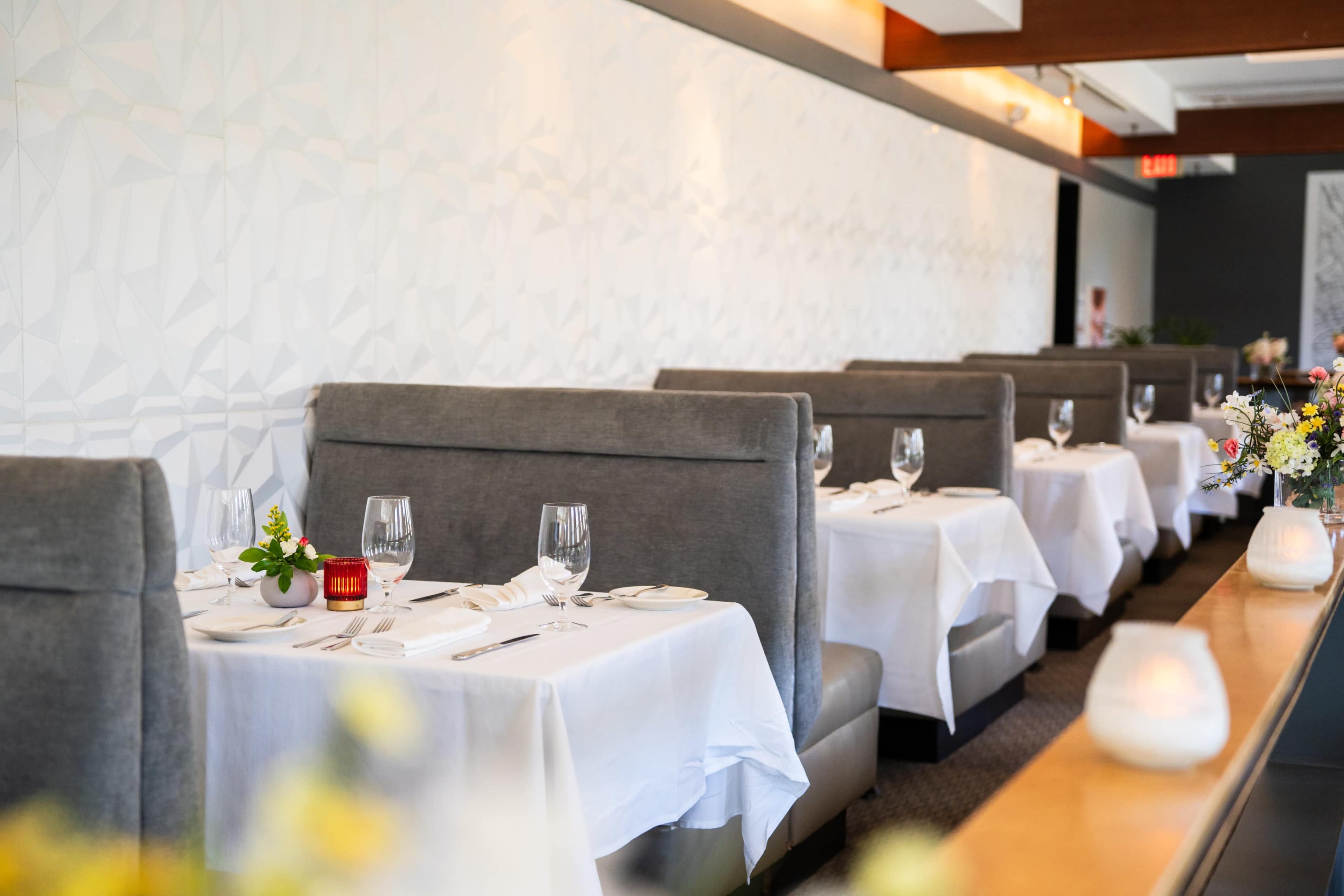 The image shows a modern restaurant interior with neatly arranged tables covered in white tablecloths, each set with glassware and cutlery.