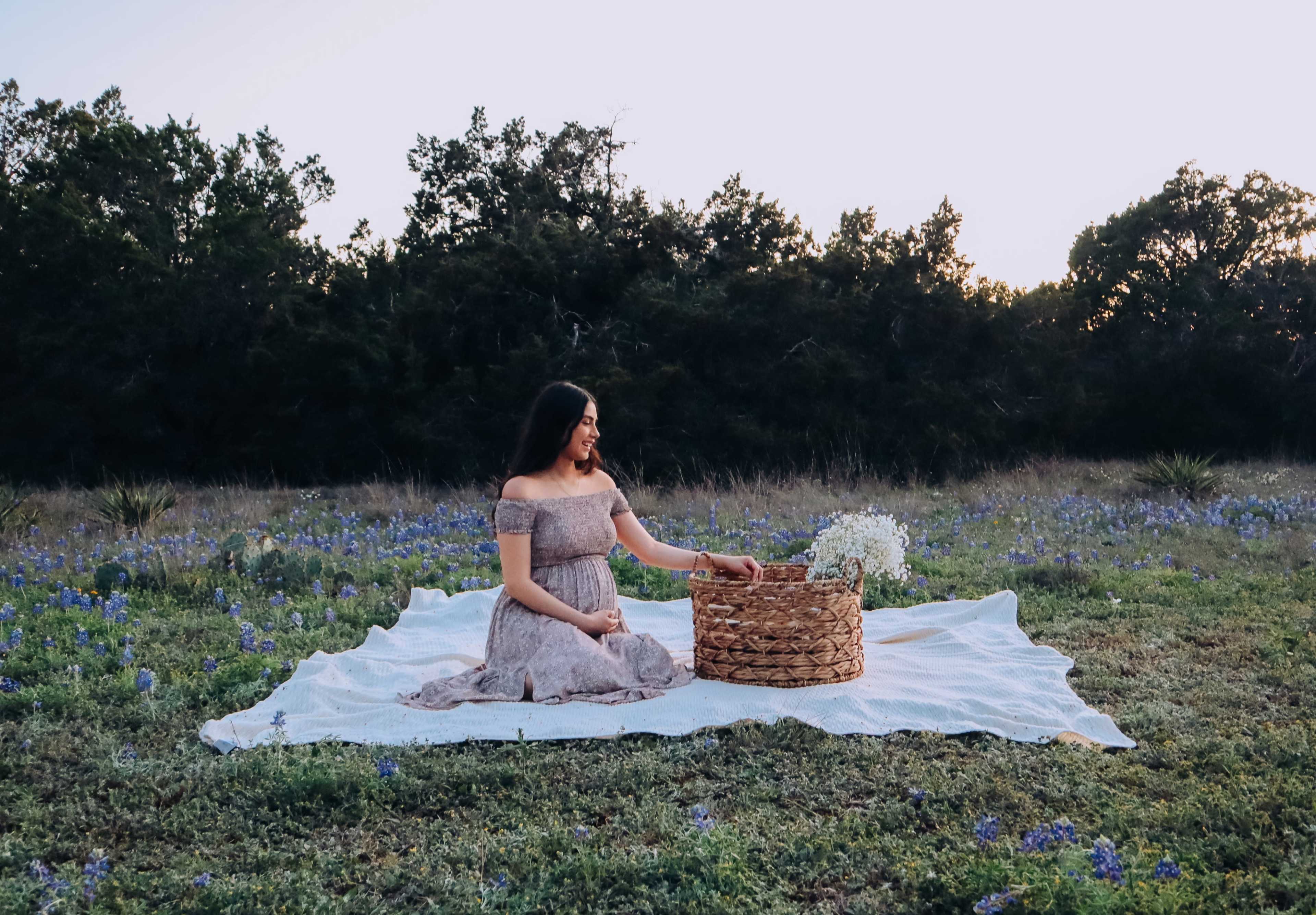 A woman sits on a blanket in a grassy field surrounded by wildflowers, next to a woven basket.