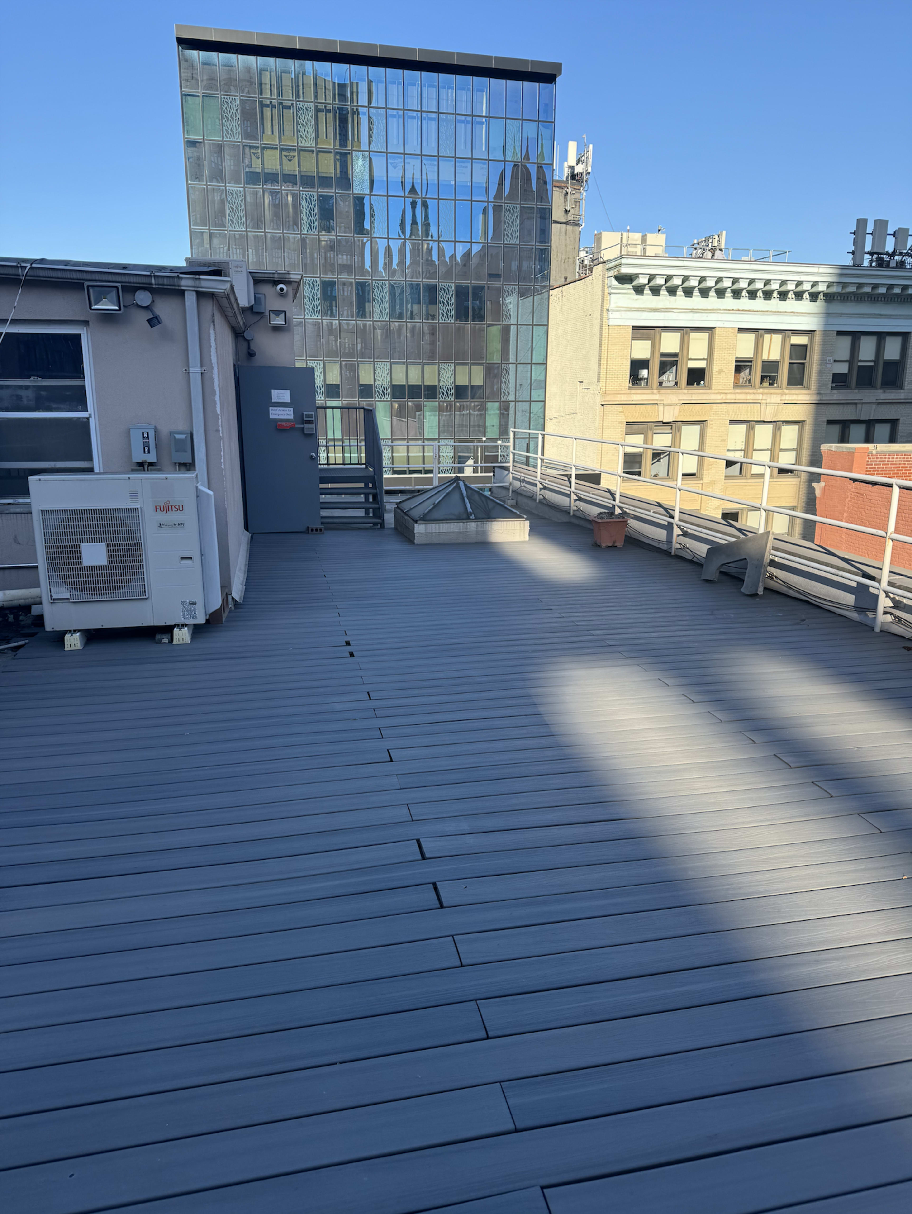 The image shows a rooftop deck with a smooth wooden surface and a glass building in the background under a clear blue sky.