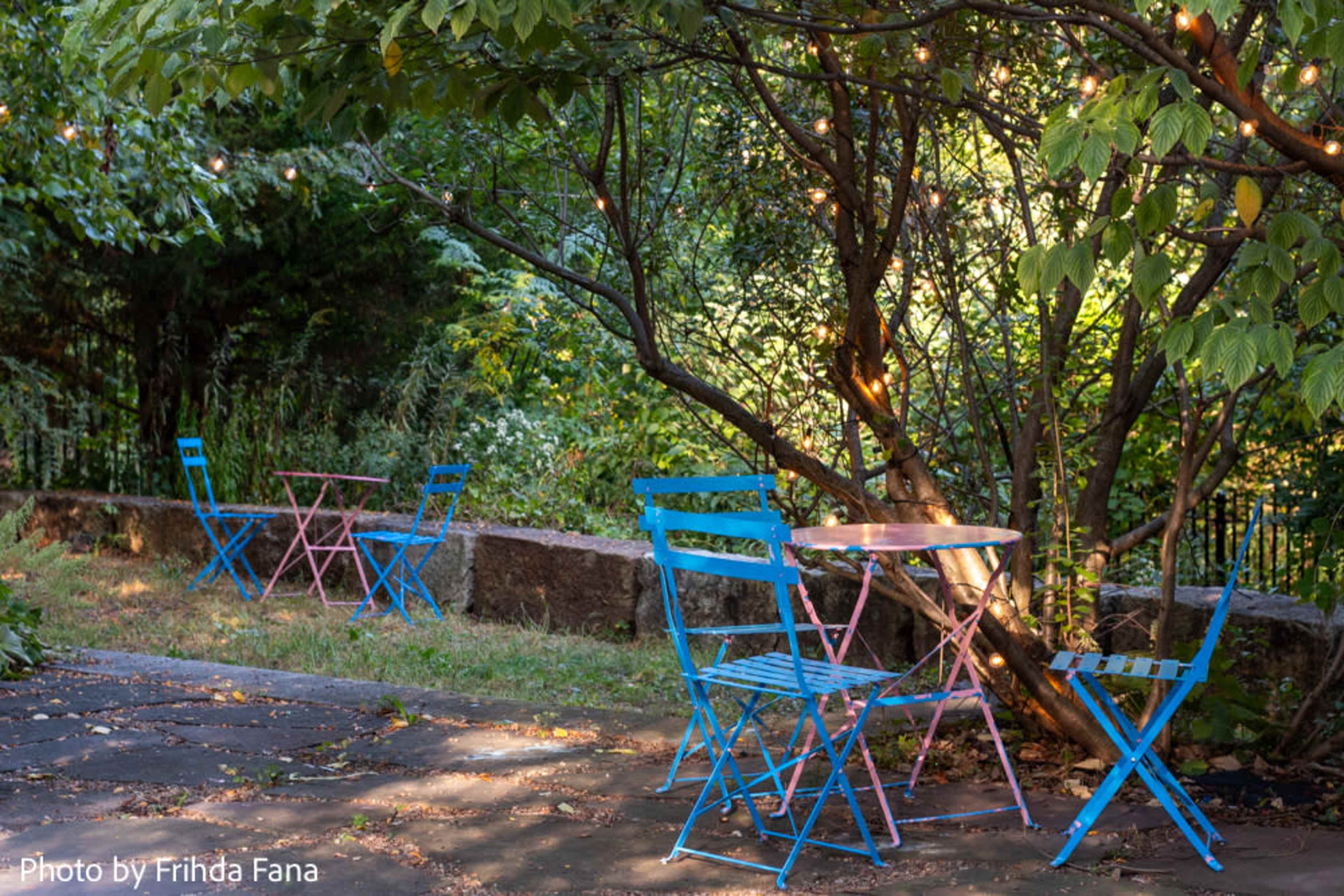 A small outdoor seating area features colorful metal chairs and a table nestled under a tree, with string lights overhead.