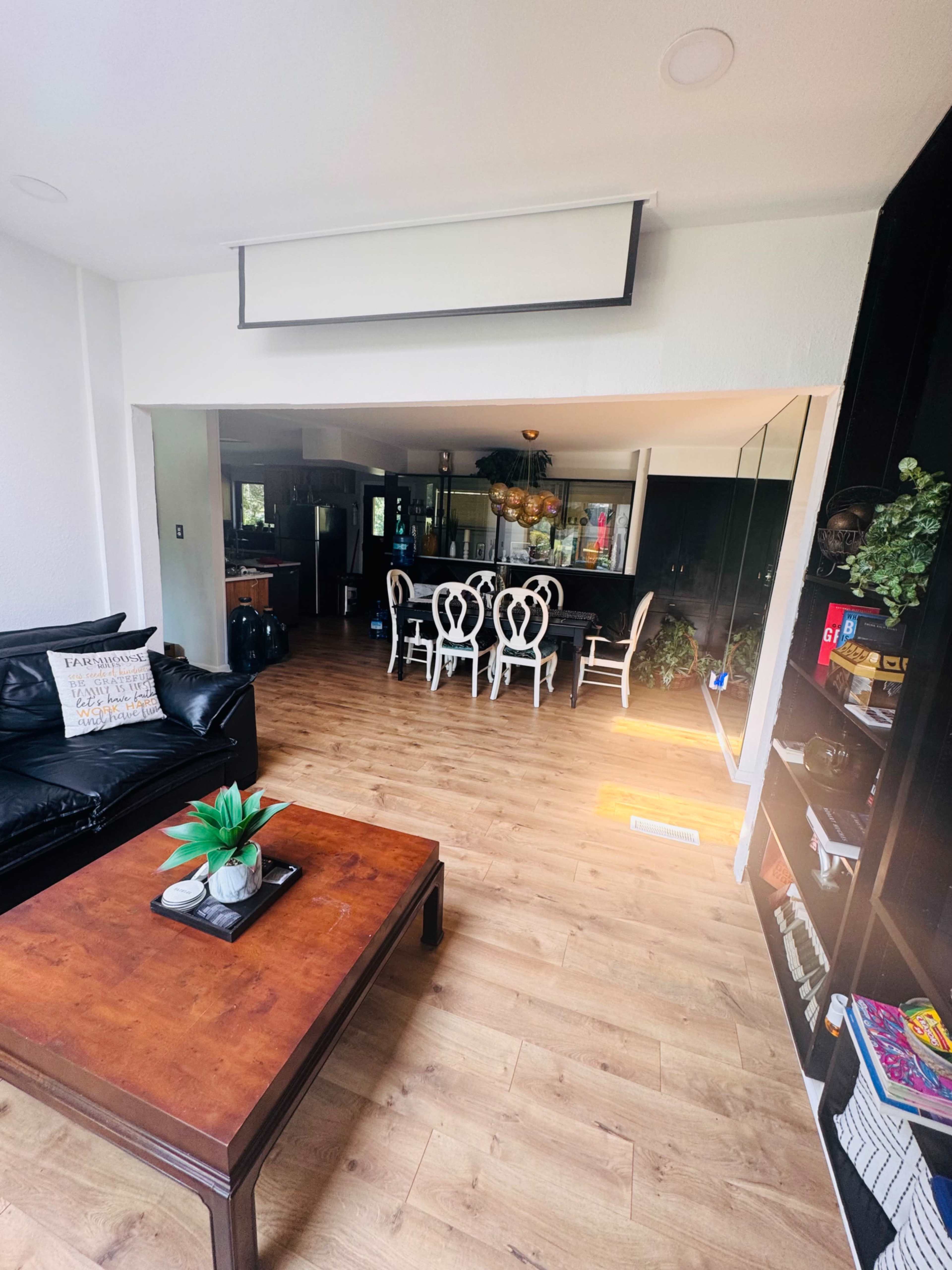 The image shows a living room with a black sofa, a wooden coffee table with a plant, and a dining area in the background featuring a table and chairs.