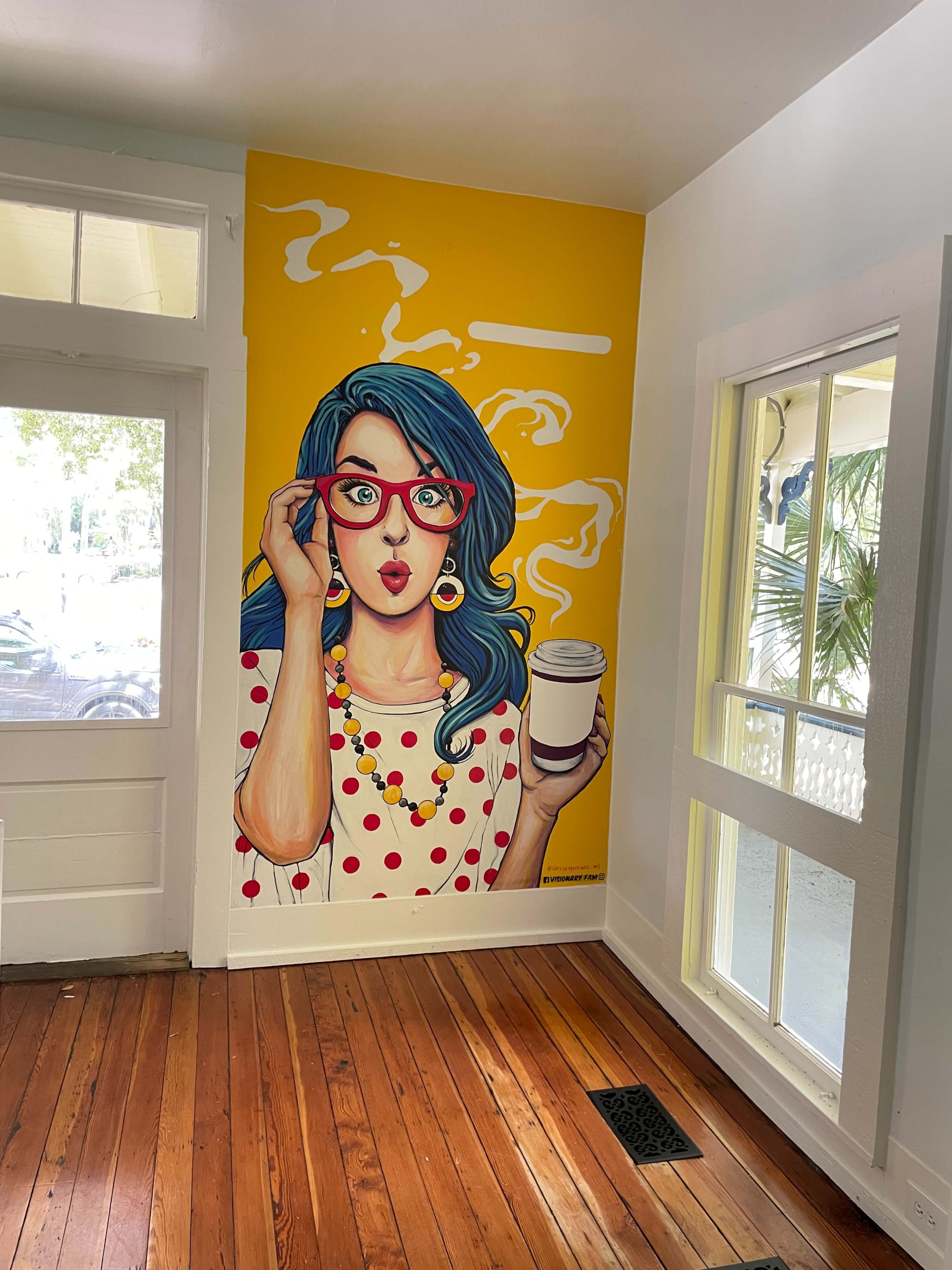 A colorful mural of a woman with blue hair and glasses holding a coffee cup adorns a wall in a brightly lit room with wooden flooring.