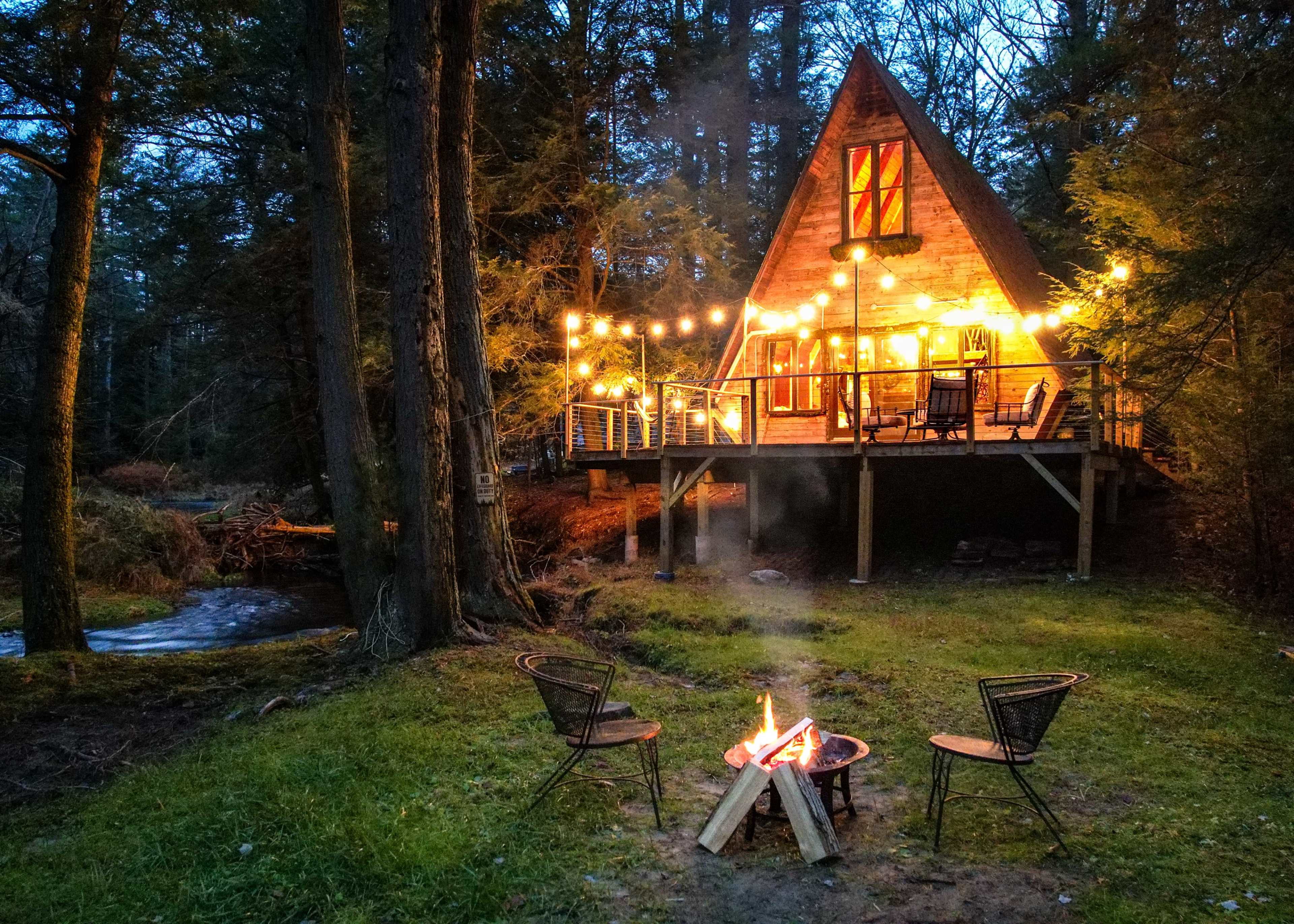 A lit A-frame cabin stands among trees, with a fire pit and three chairs arranged in front of it near a flowing stream.