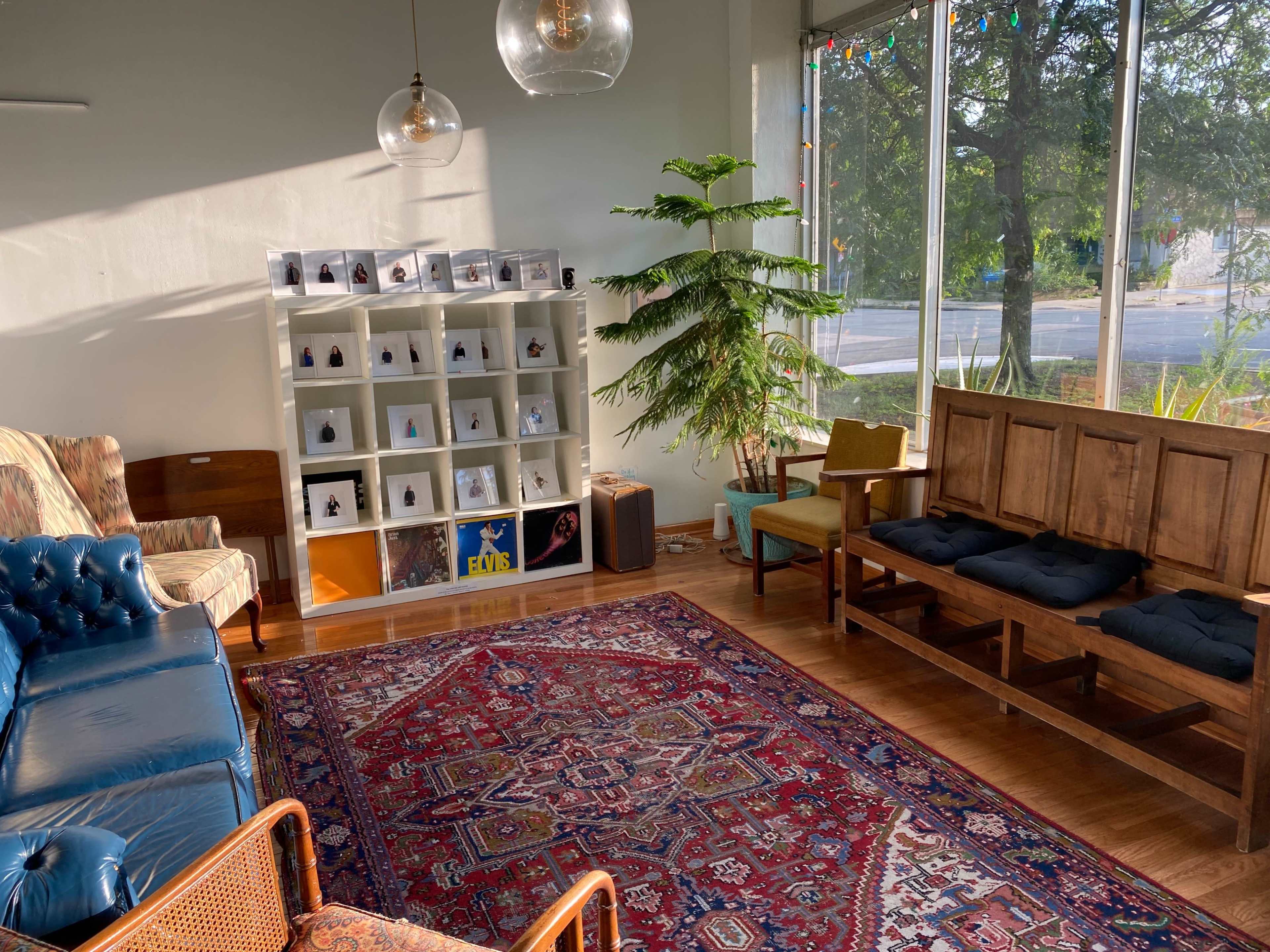 The image shows a cozy room with a blue leather sofa, a wooden seating area, a bookshelf displaying small objects, and a large window letting in natural light.