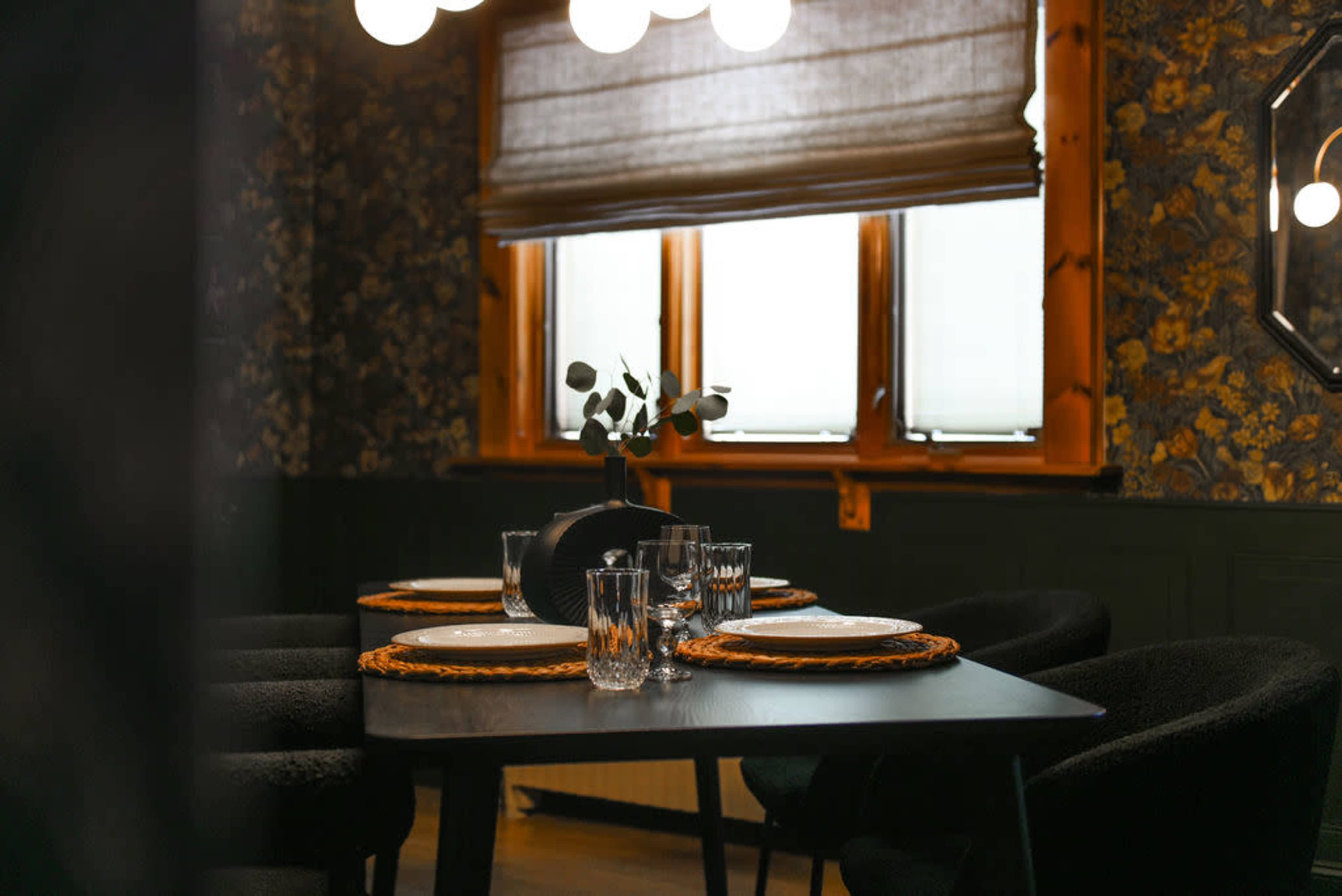 A dining area features a table set with plates and glassware, illuminated by pendant lights, against a backdrop of floral wallpaper and wooden window frames.