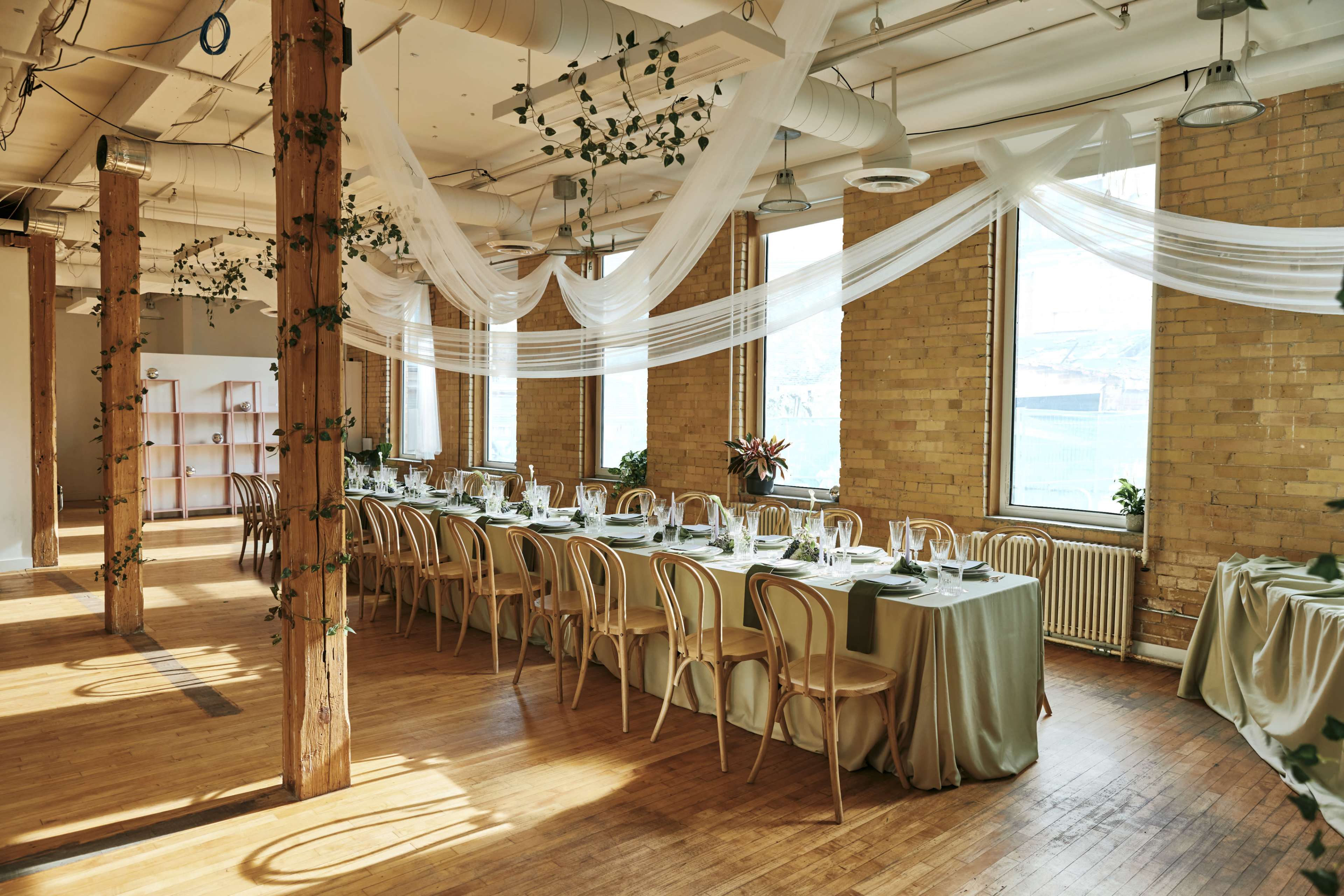 The image shows a dining setup in a spacious room with wooden beams, featuring a long table elegantly arranged with tableware and decorated with greenery, under flowing fabric drapes.