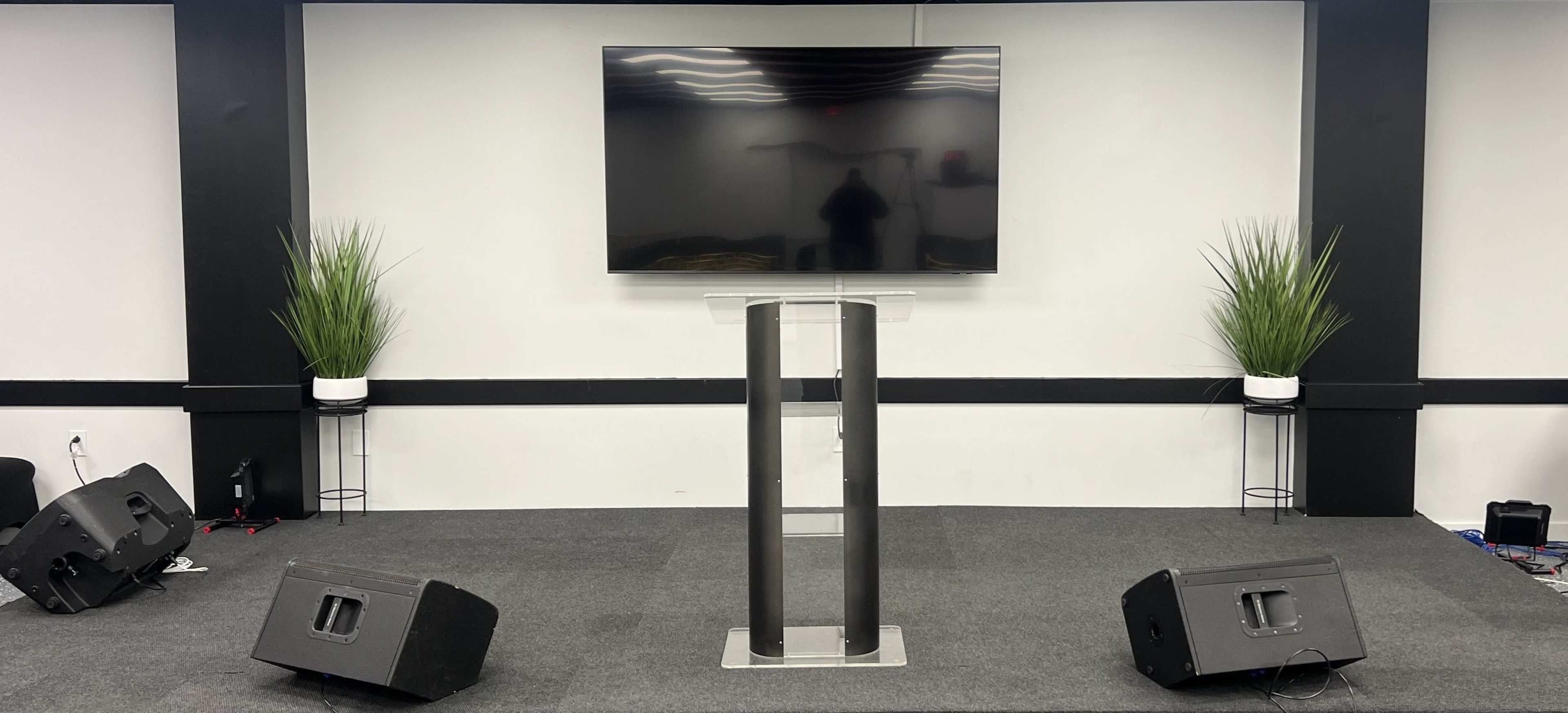The image shows a minimalist stage with a flat-screen TV mounted above a clear podium, flanked by potted plants and two speakers on either side.