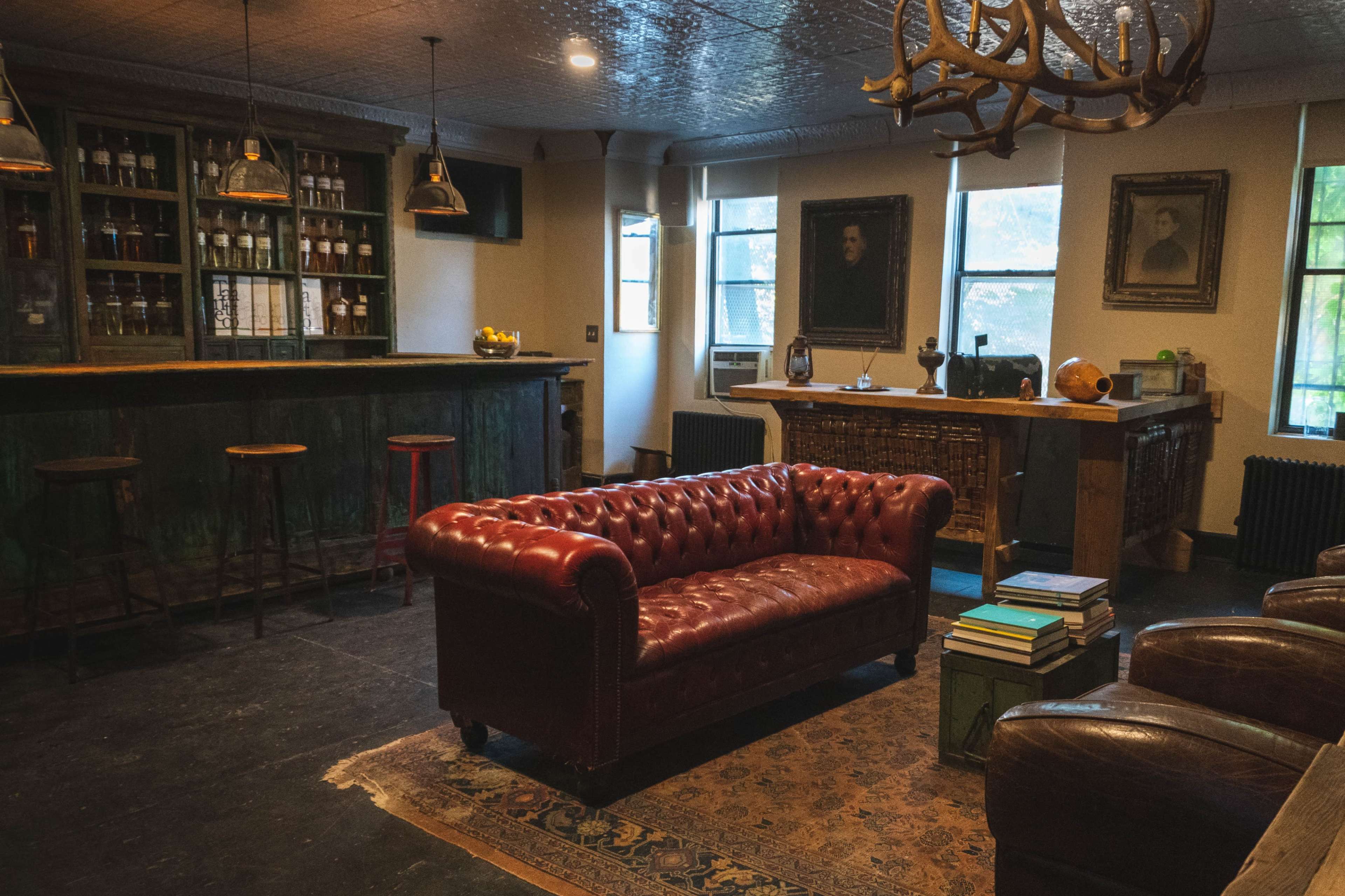 A cozy lounge area featuring a leather Chesterfield sofa, a wooden bar with shelves filled with bottles, and a mix of antique furniture.