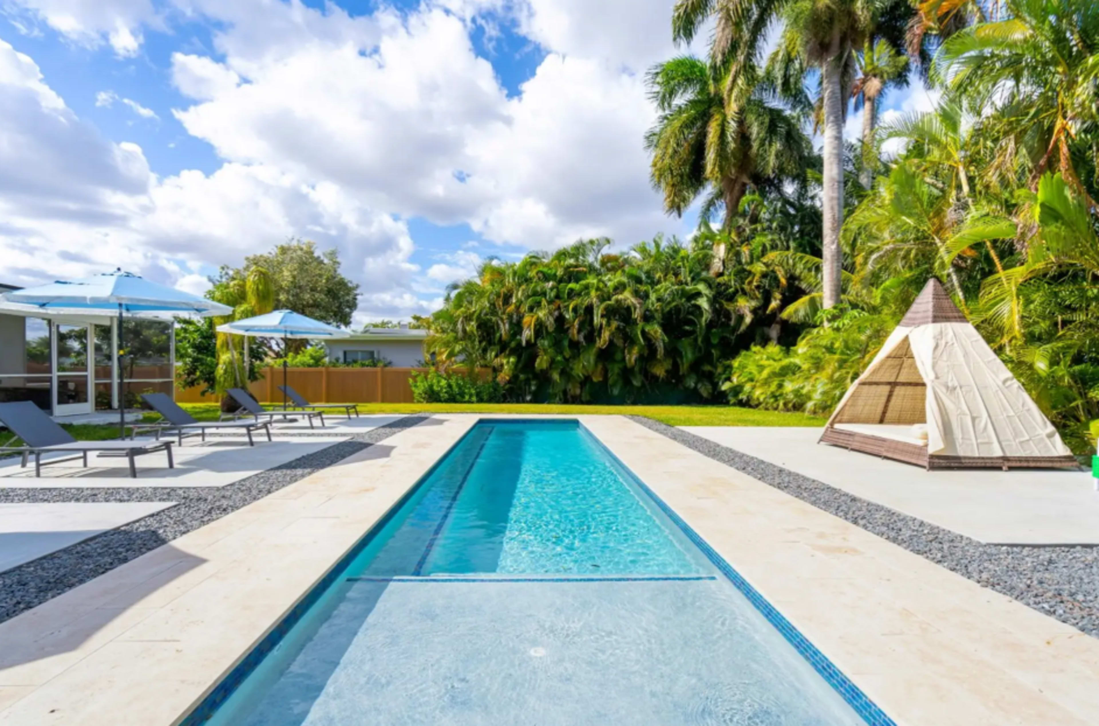 The image shows a modern backyard with a long swimming pool bordered by lounge chairs and umbrellas, surrounded by greenery and palm trees.