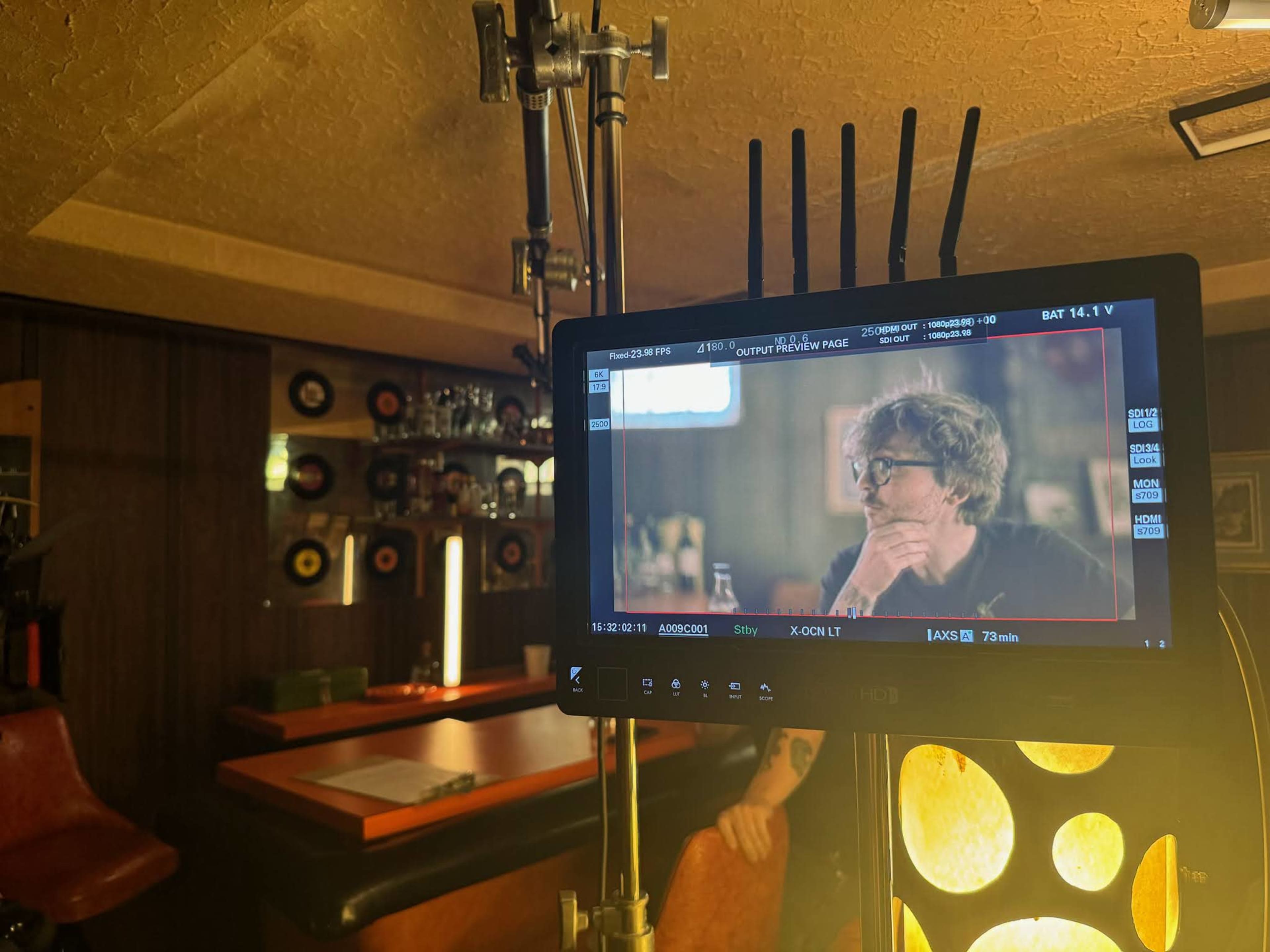 A film or television crew is set up in a bar, with a monitor displaying a scene featuring a man with glasses sitting at a table.