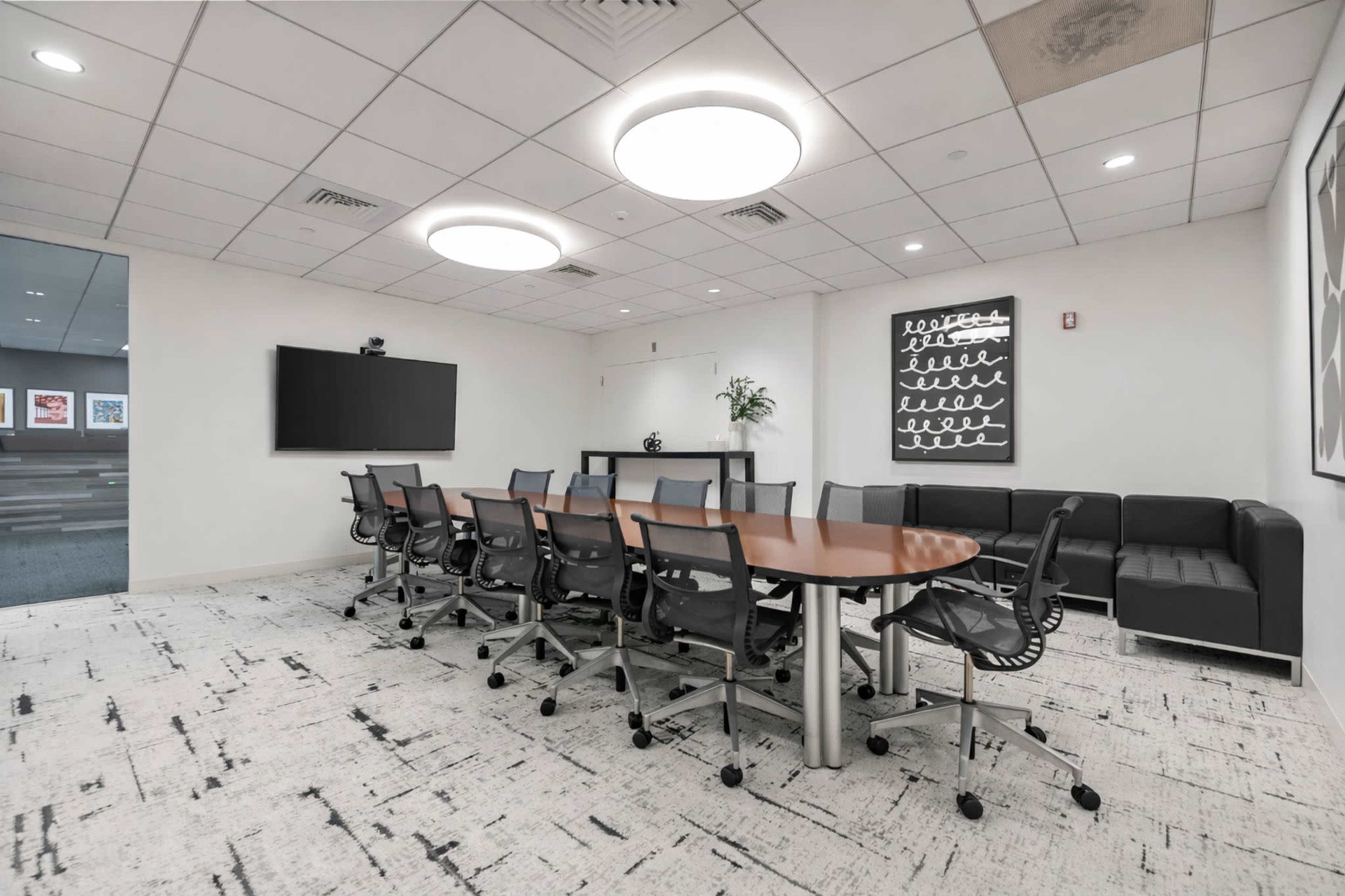 A modern conference room features a large wooden table surrounded by black office chairs, with a couch and a wall-mounted TV in the background.
