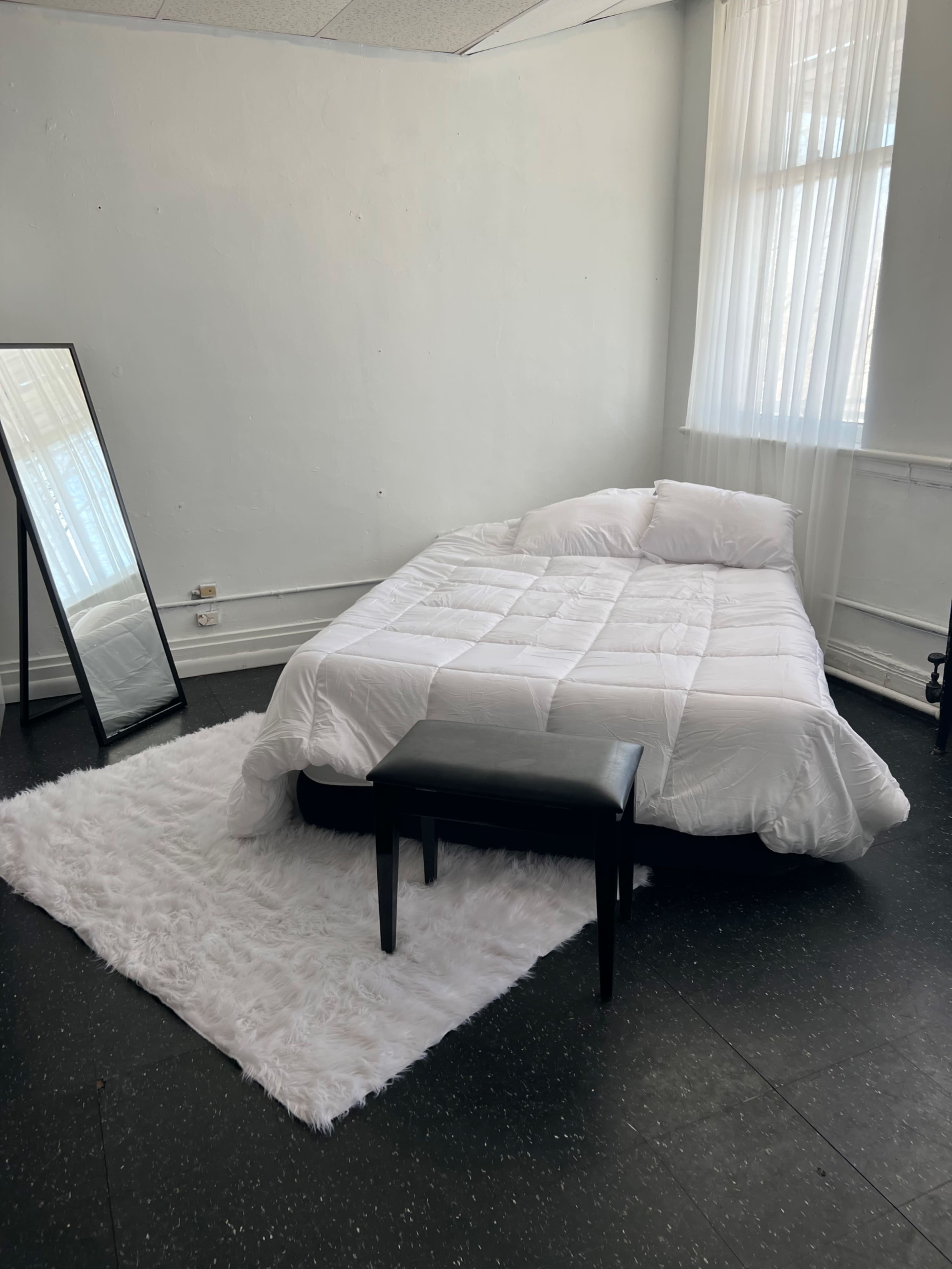 A simple bedroom with a bed covered in a white comforter, a black stool in front of it, a large mirror, and a fluffy white rug on the floor.