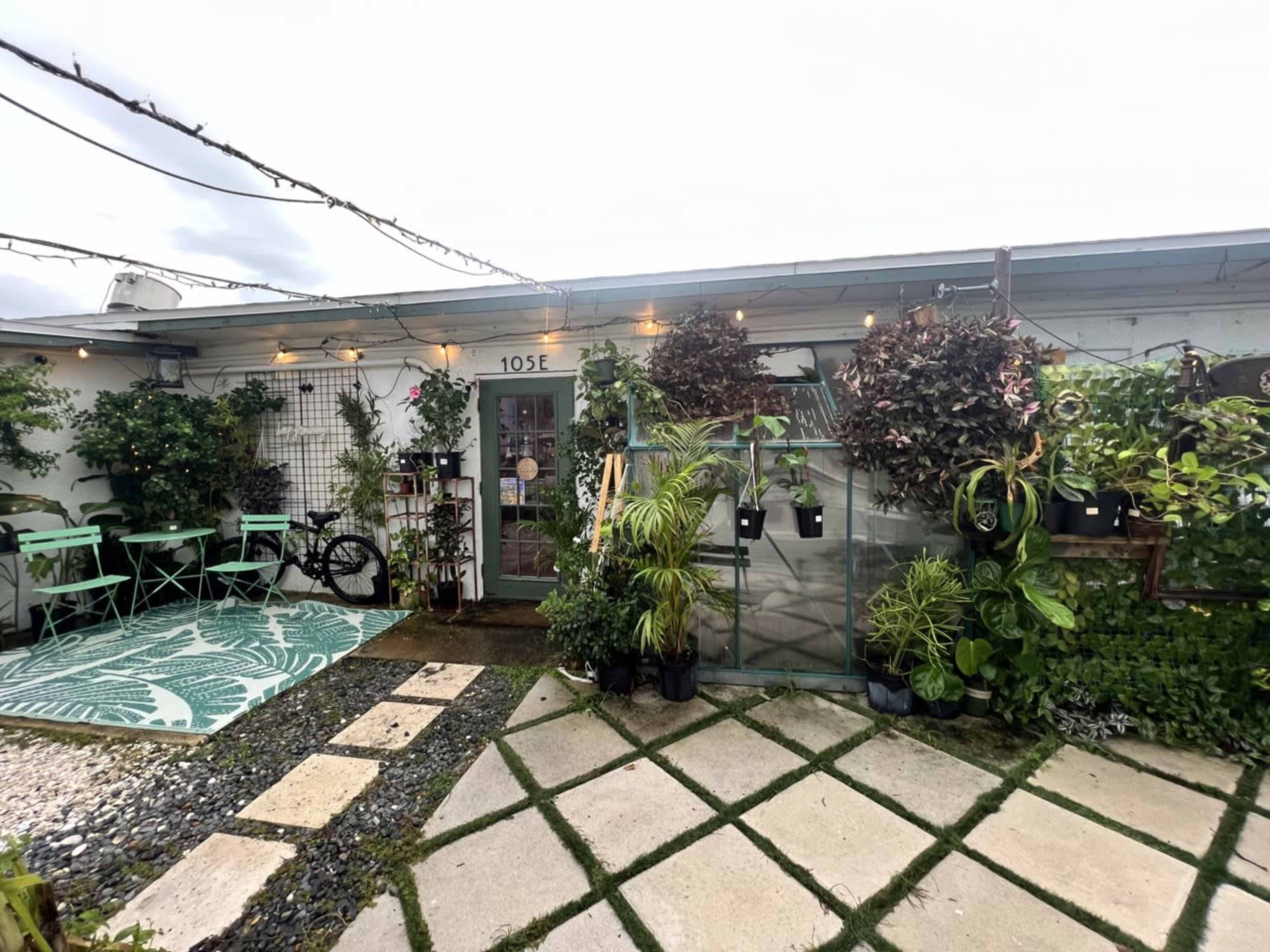 The image shows a small outdoor garden area with various potted plants, a patterned rug, two green chairs, and a decorative entrance.