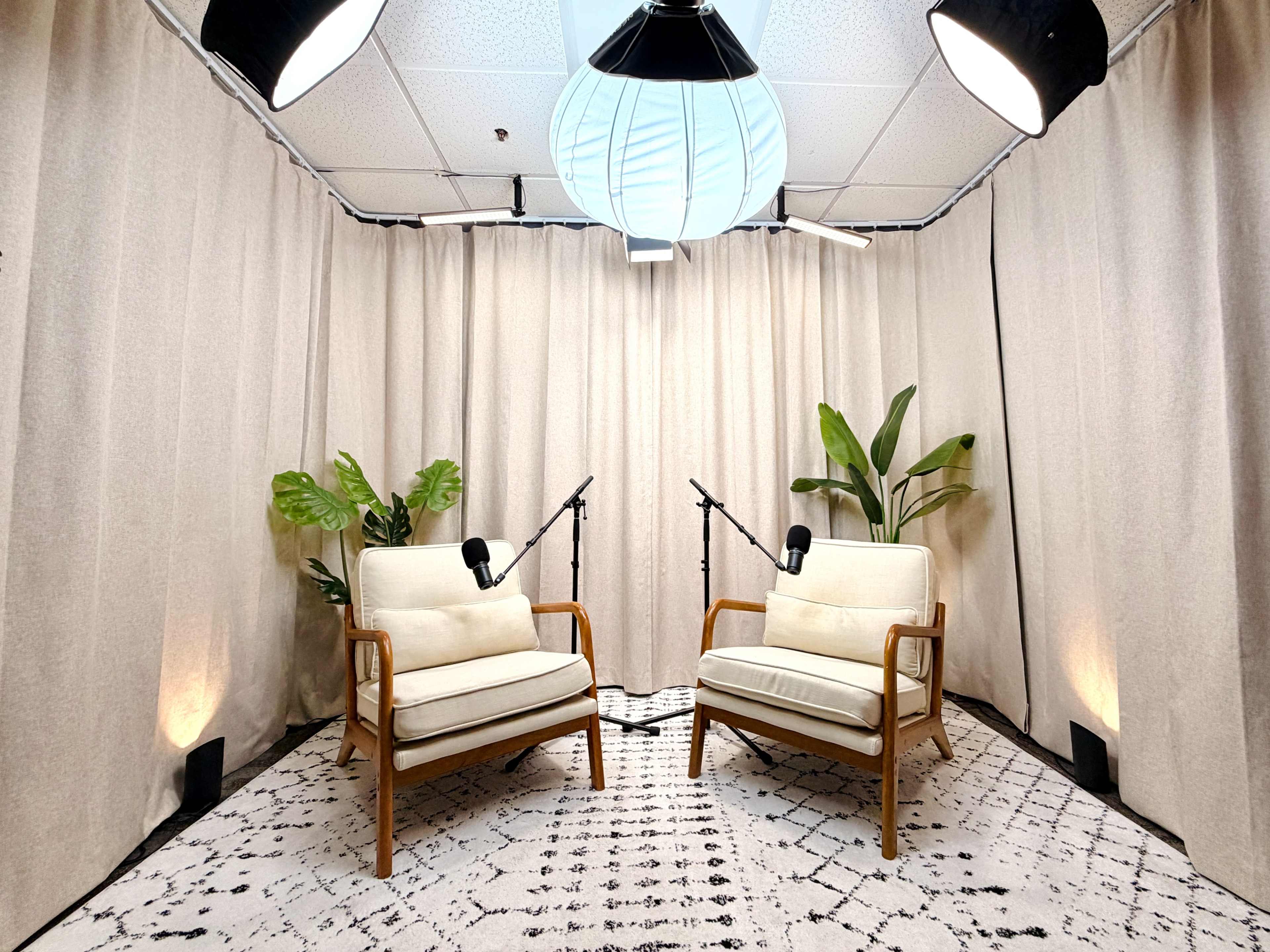The image shows a small studio setup with two wooden-framed armchairs facing each other, surrounded by fabric curtains, plants, and microphones on stands.