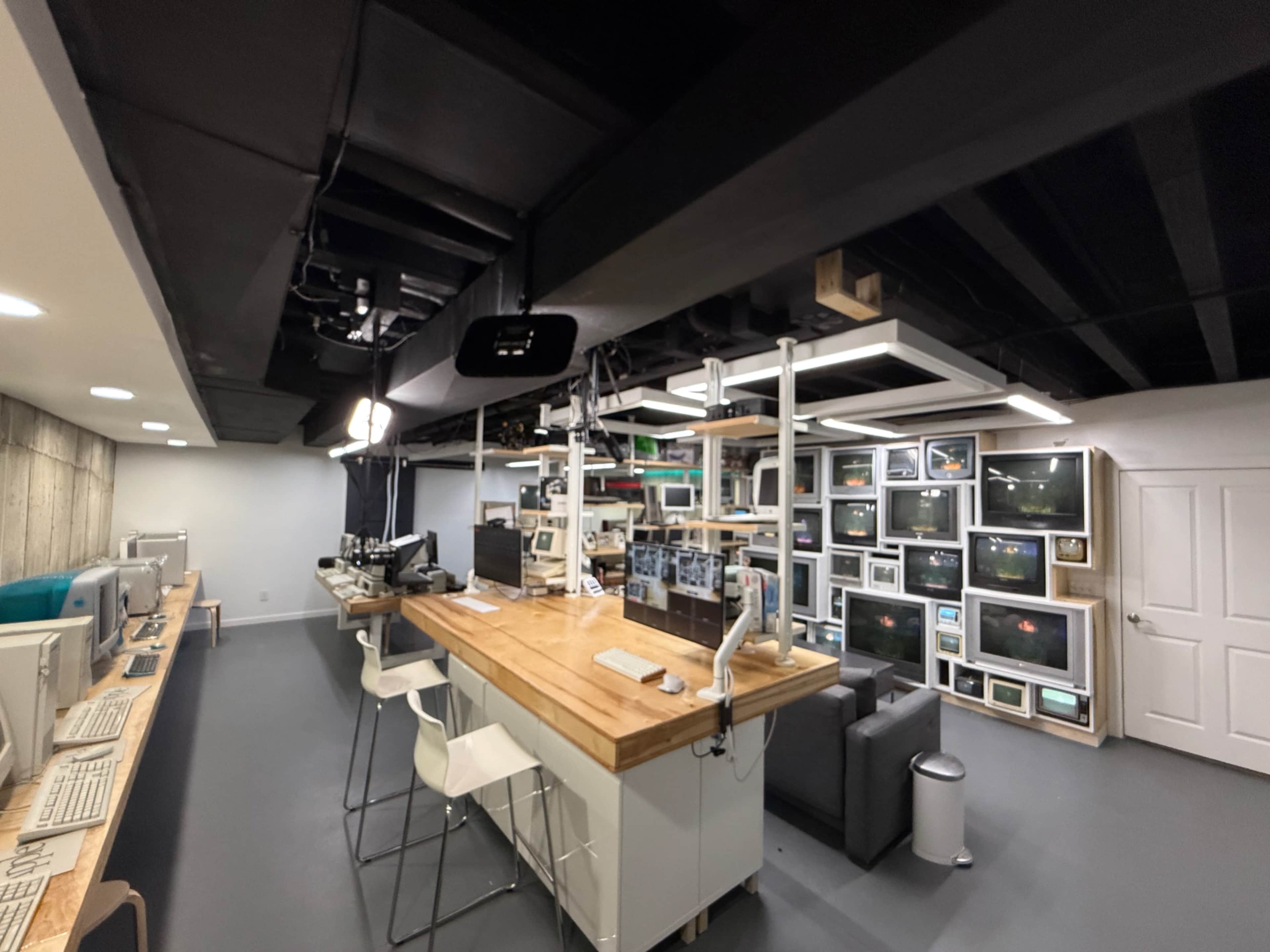 The image shows a modern basement workspace featuring multiple vintage computers, a large wooden island, and several old television sets displayed against the wall.