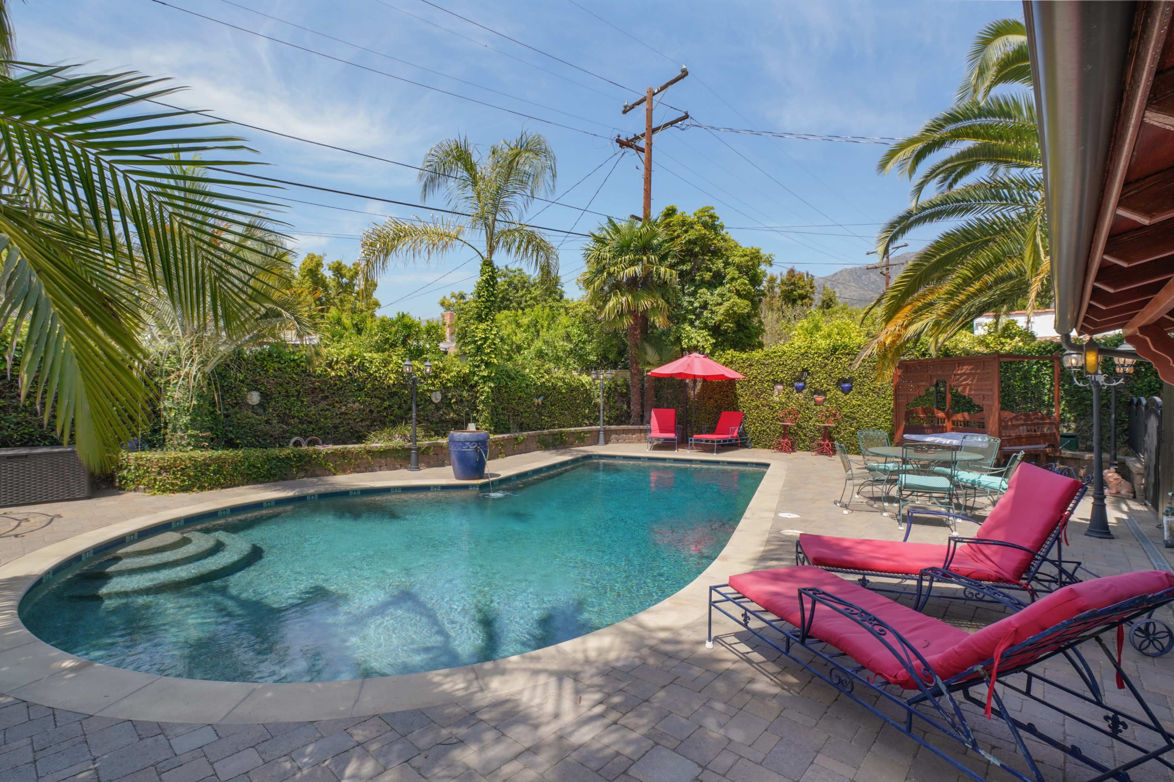 A backyard features a swimming pool surrounded by lounge chairs, potted plants, and a red umbrella under a clear blue sky.