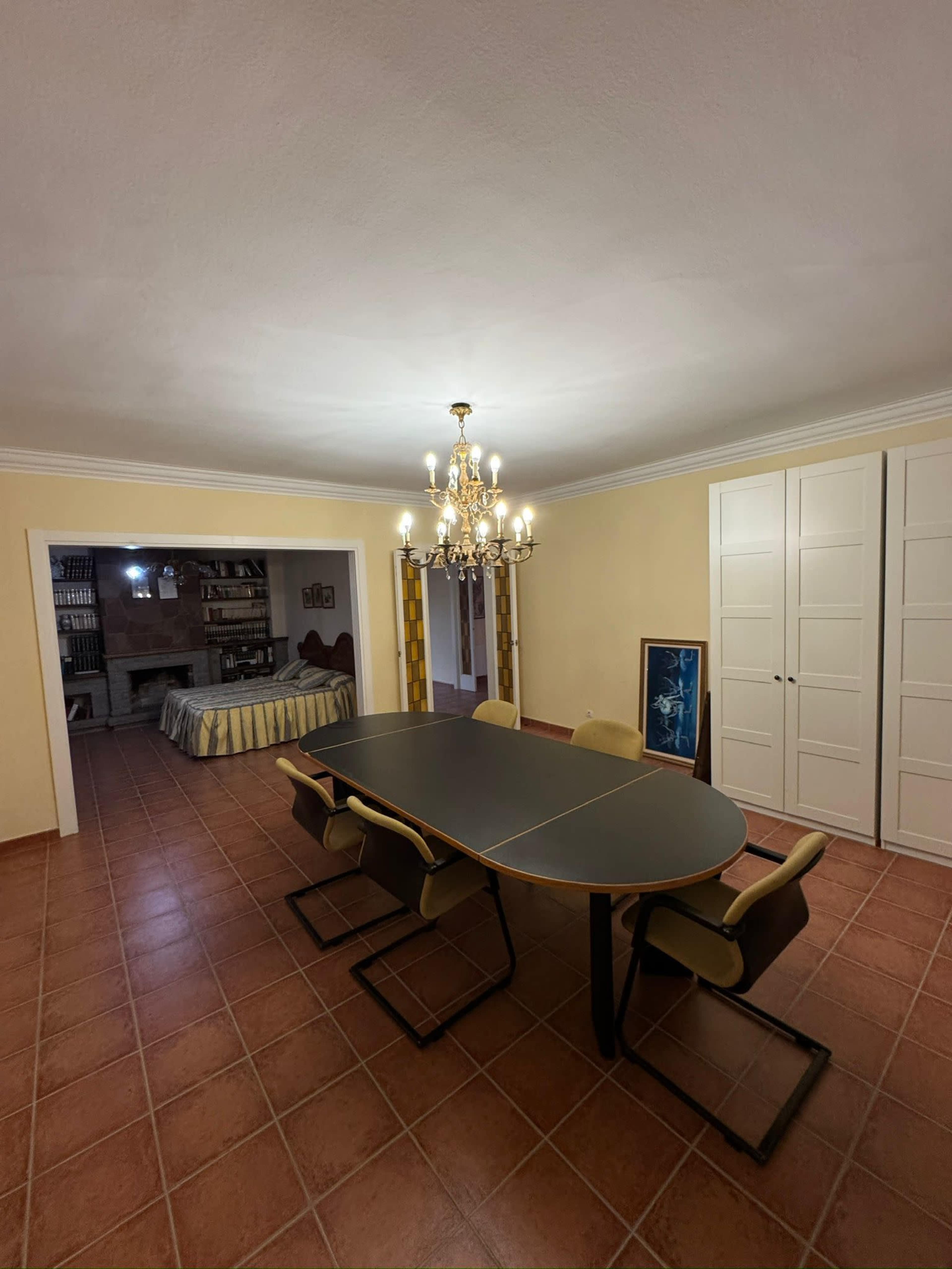The image shows a dining area with a large table and chairs, a chandelier overhead, and an open doorway leading to another room with a bed visible in the background.