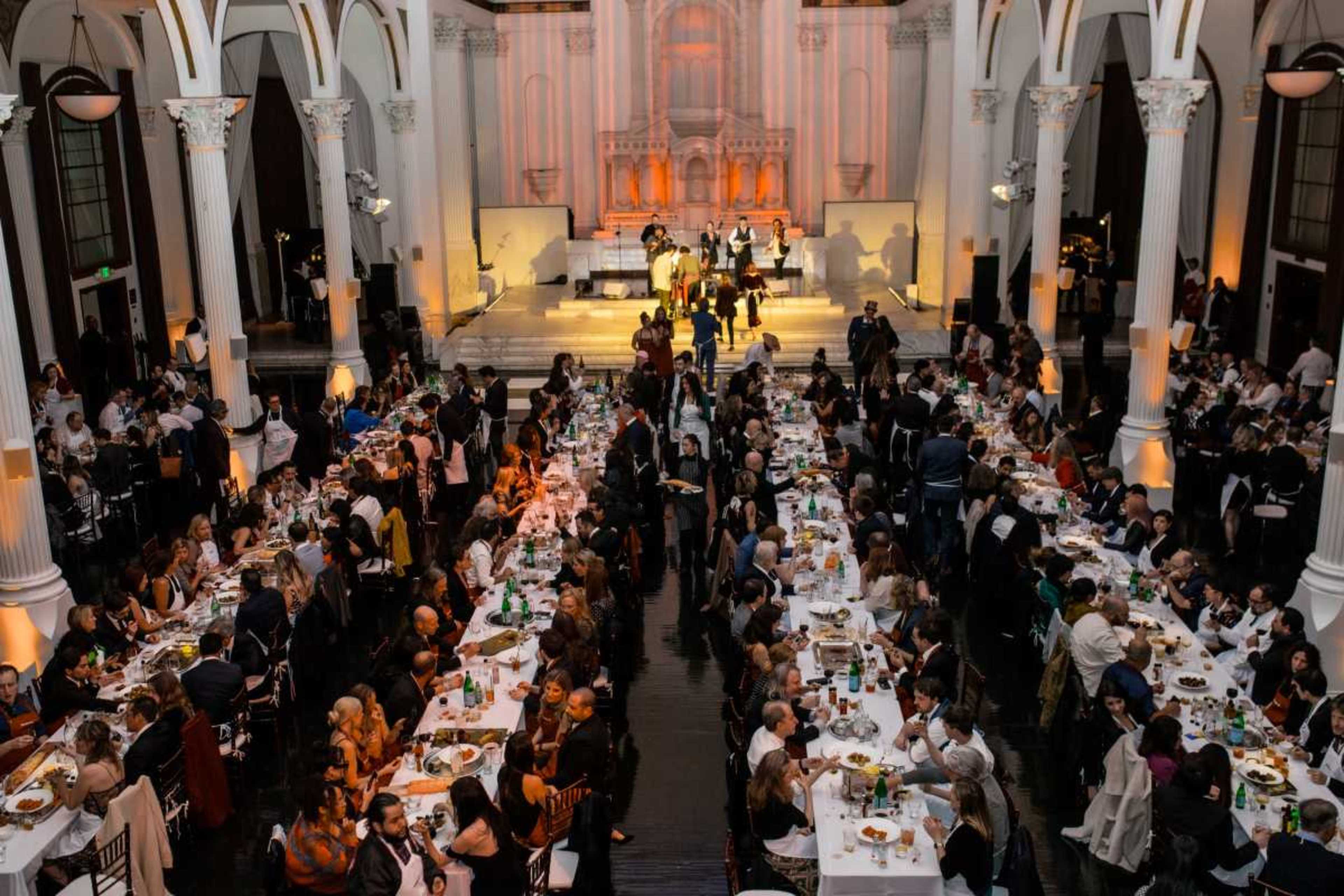A large crowd attends a formal dinner event in a spacious hall adorned with tall columns and dramatic lighting.
