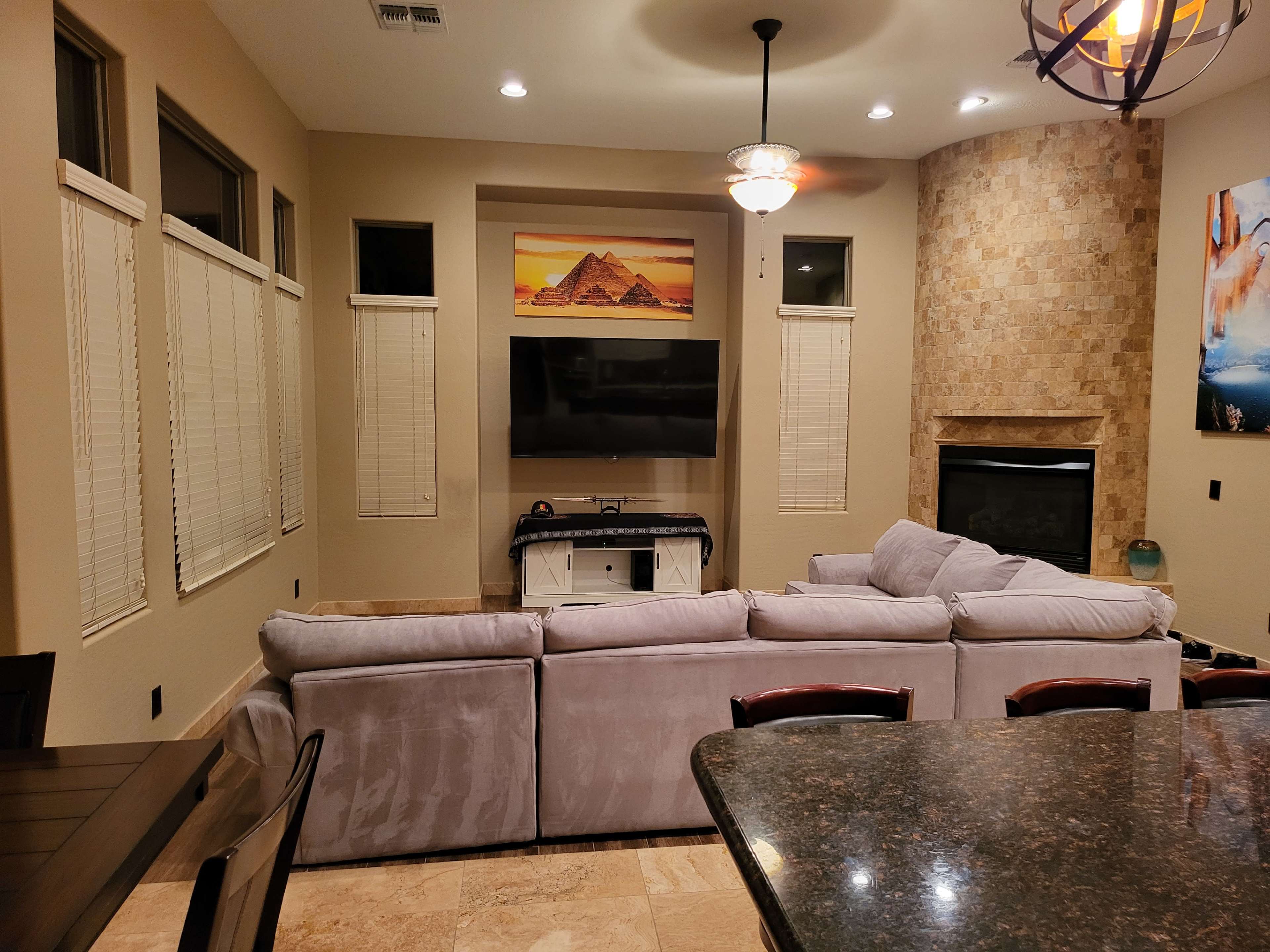 A living room with a sectional sofa, a television mounted on the wall, and a stone fireplace, all set in a neutral-toned environment with window shades.