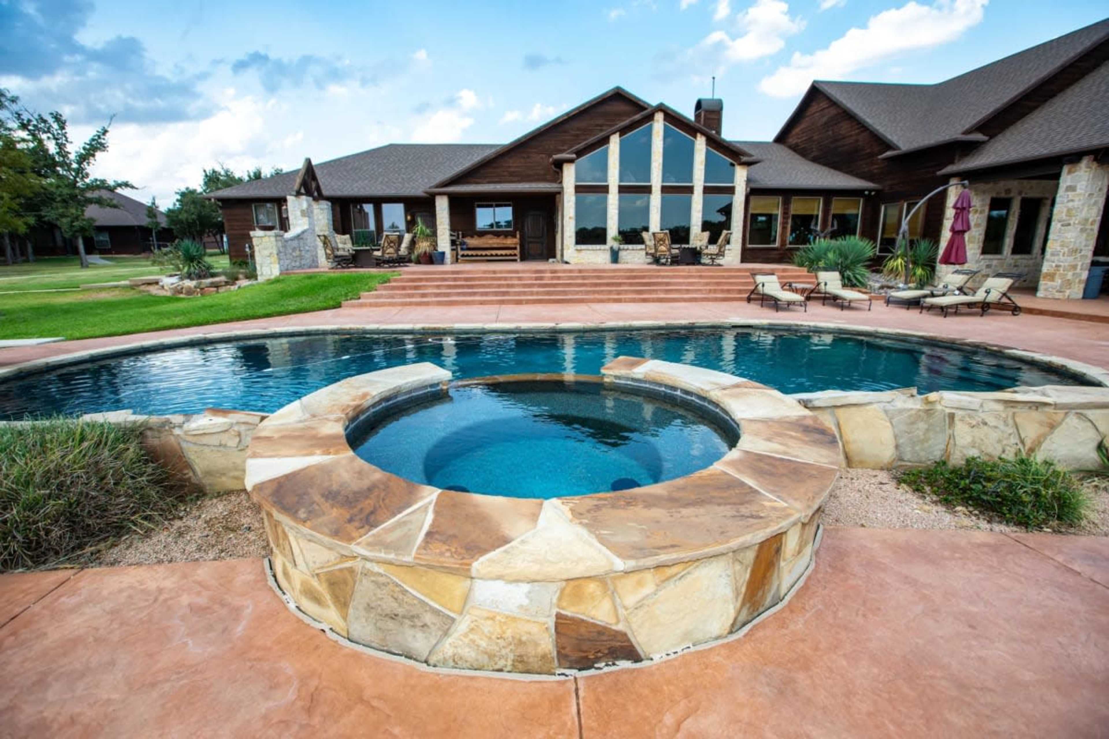 The image shows a modern outdoor pool area with a circular hot tub, surrounded by stone and concrete, and a spacious house in the background.