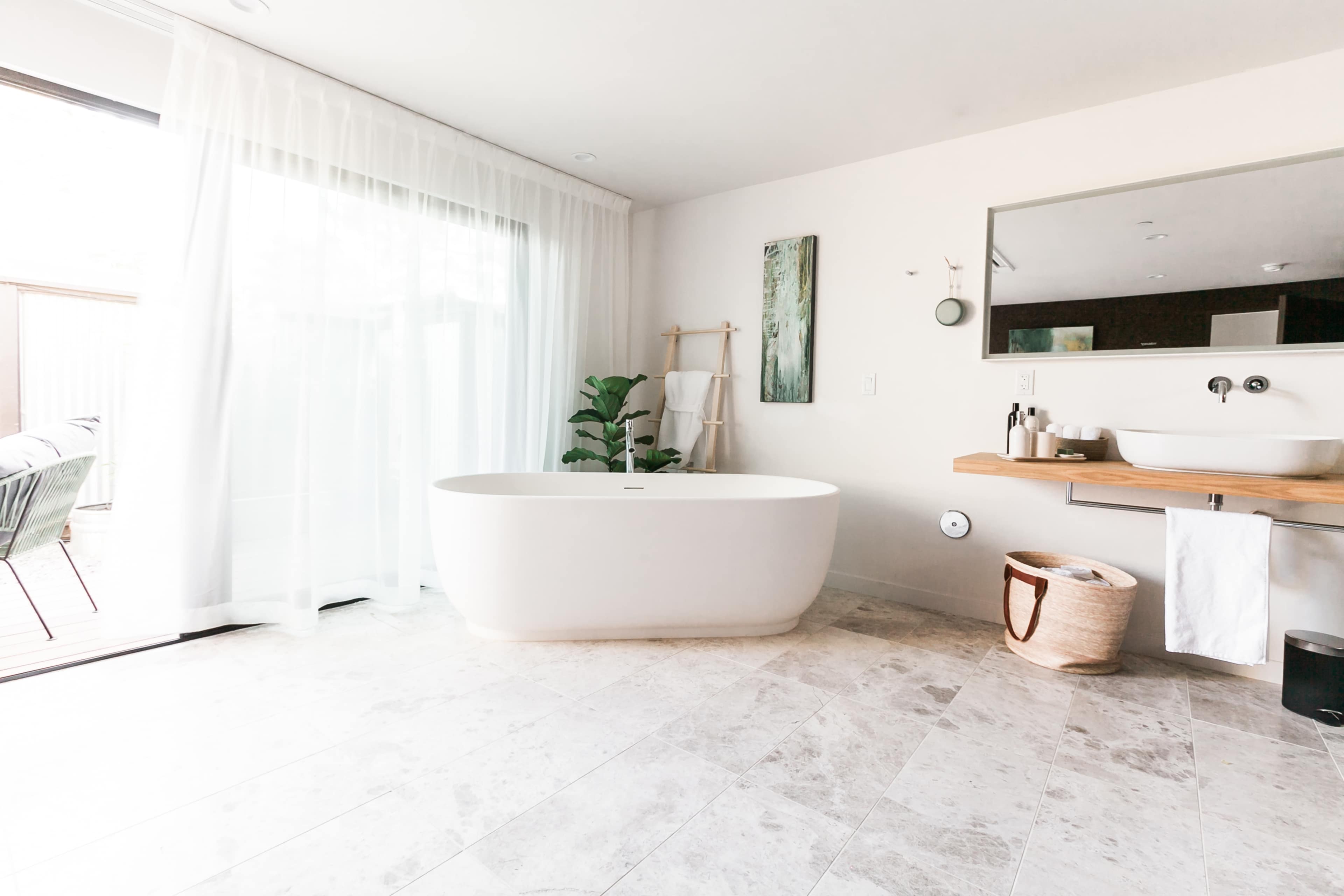 The image shows a modern bathroom with a freestanding bathtub, a wooden counter with a sink, and large windows allowing natural light.