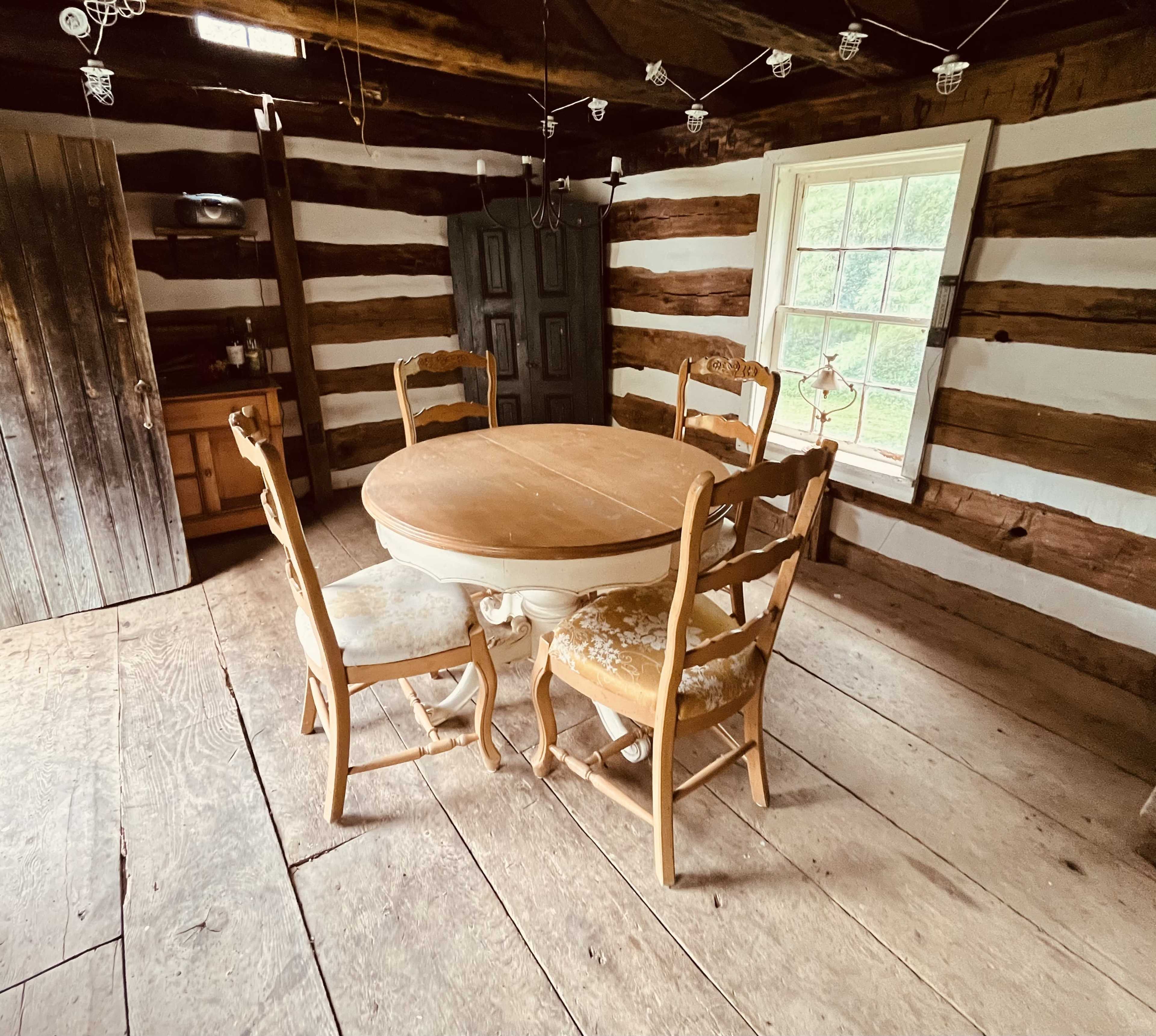 The image shows a rustic room with exposed wooden beams, featuring a round wooden table surrounded by four chairs, and a window letting in natural light.