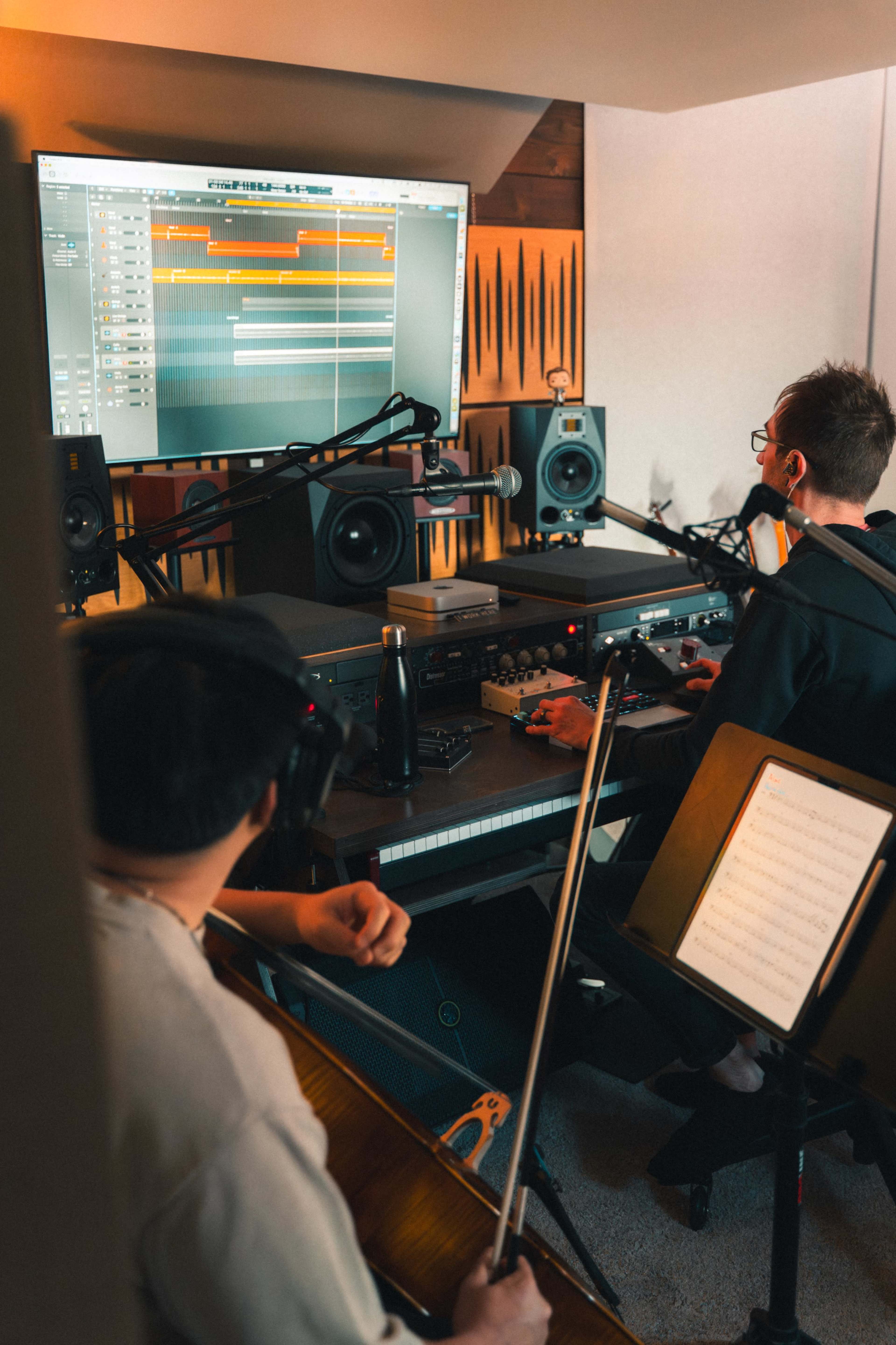 Two musicians collaborate in a recording studio, with one playing a violin and the other mixing tracks on a computer while surrounded by audio equipment.