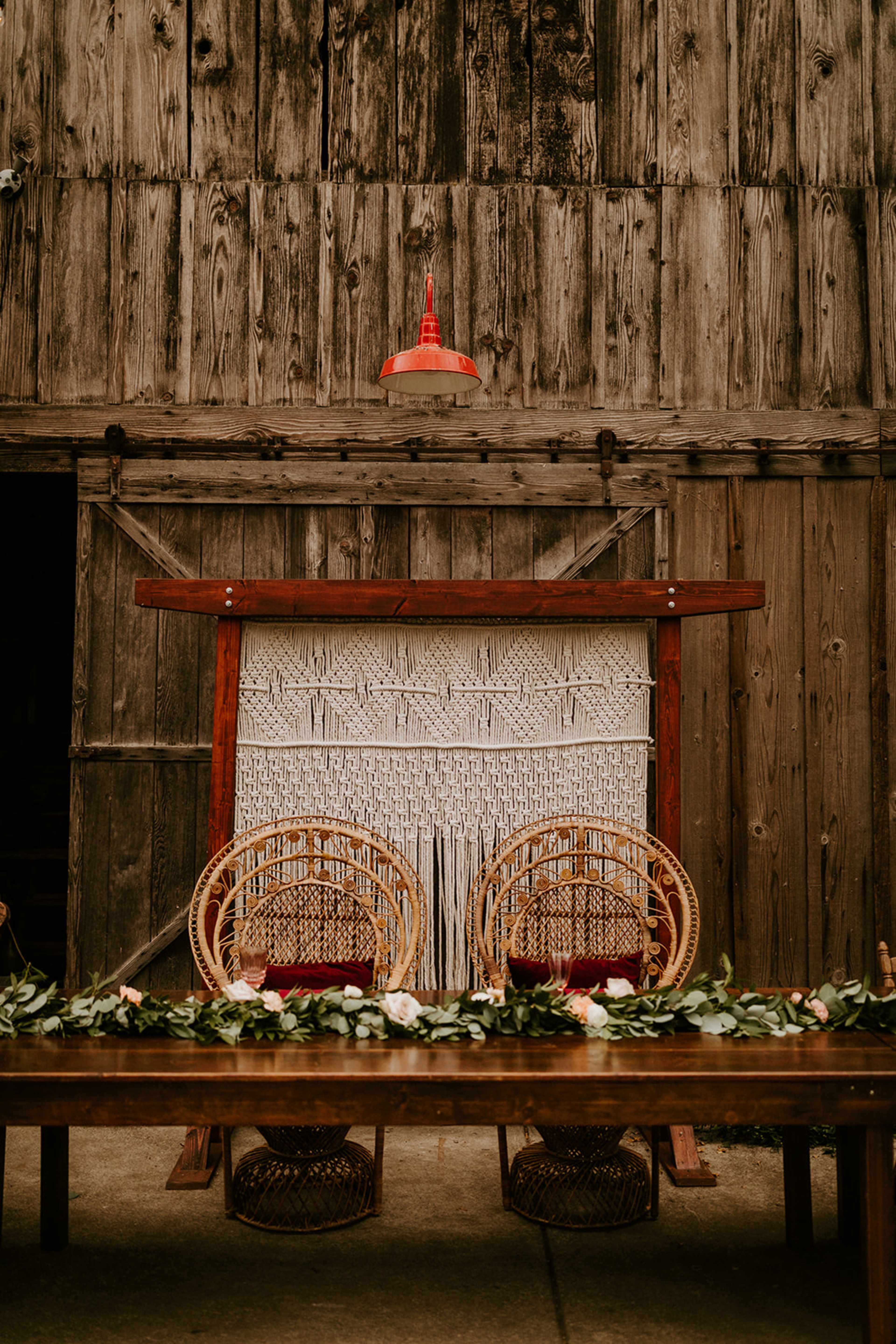 A wooden table with two rattan chairs is set against a rustic barn wall, featuring a macramé backdrop and greenery along the table's edge.