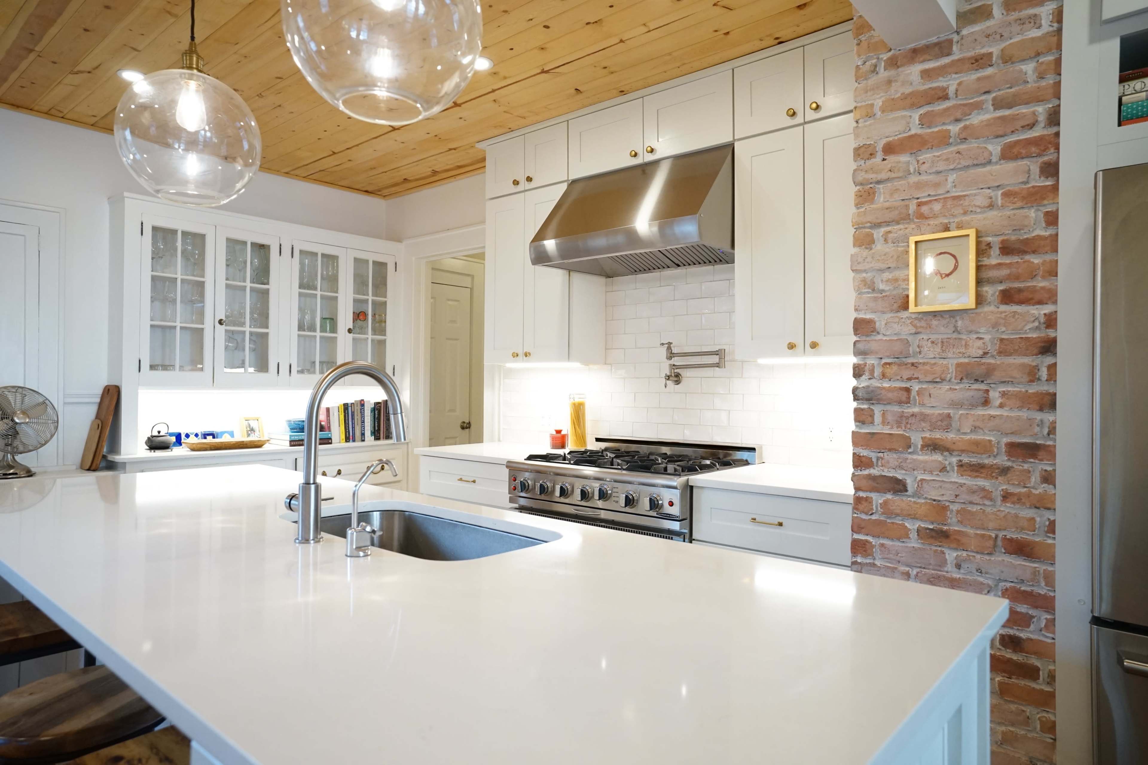 A modern kitchen features a white countertop, stainless steel appliances, brick walls, and wooden ceiling beams.
