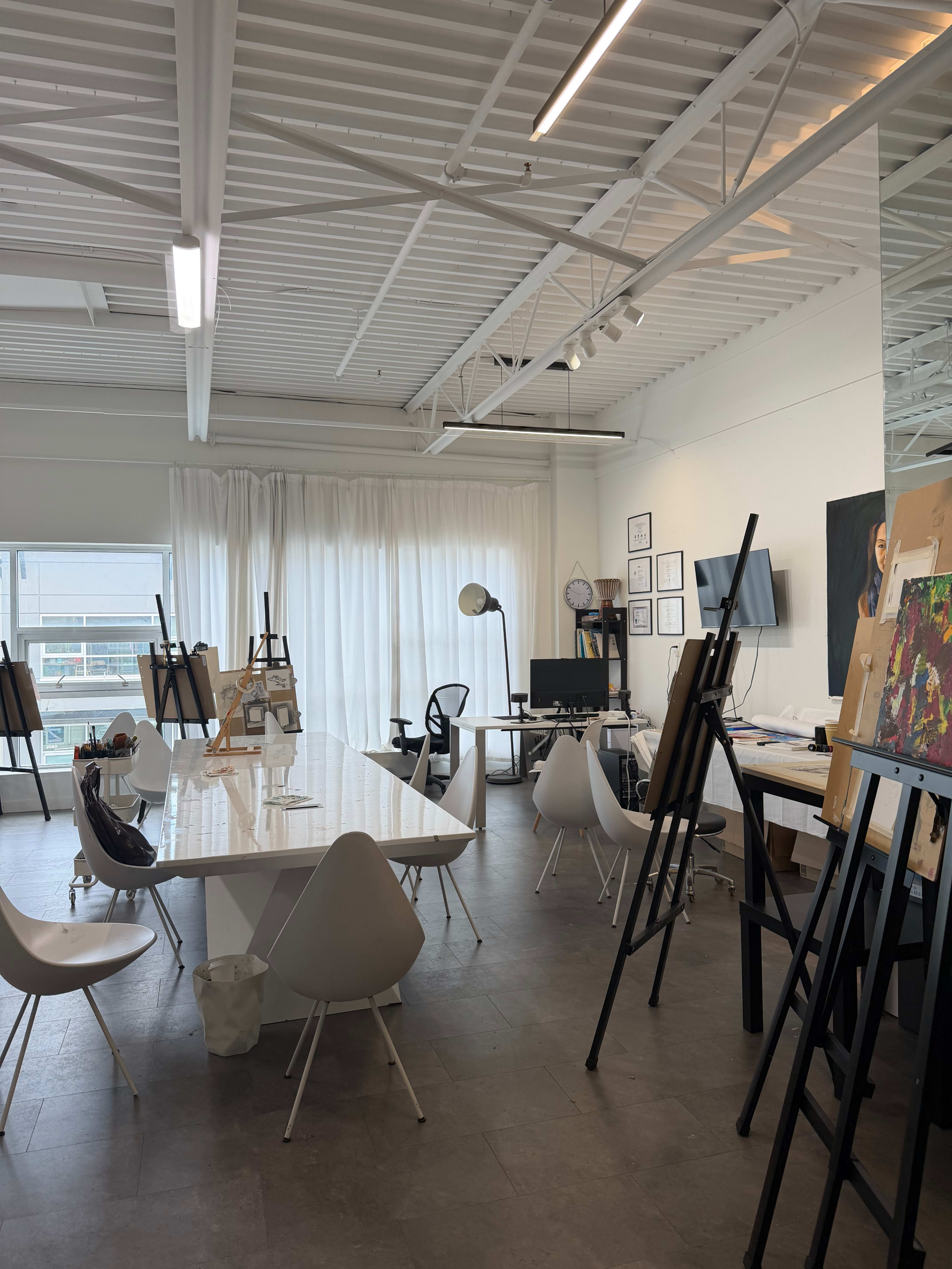 The image shows a well-lit art studio featuring easels, a large table, and various pieces of art on the walls.