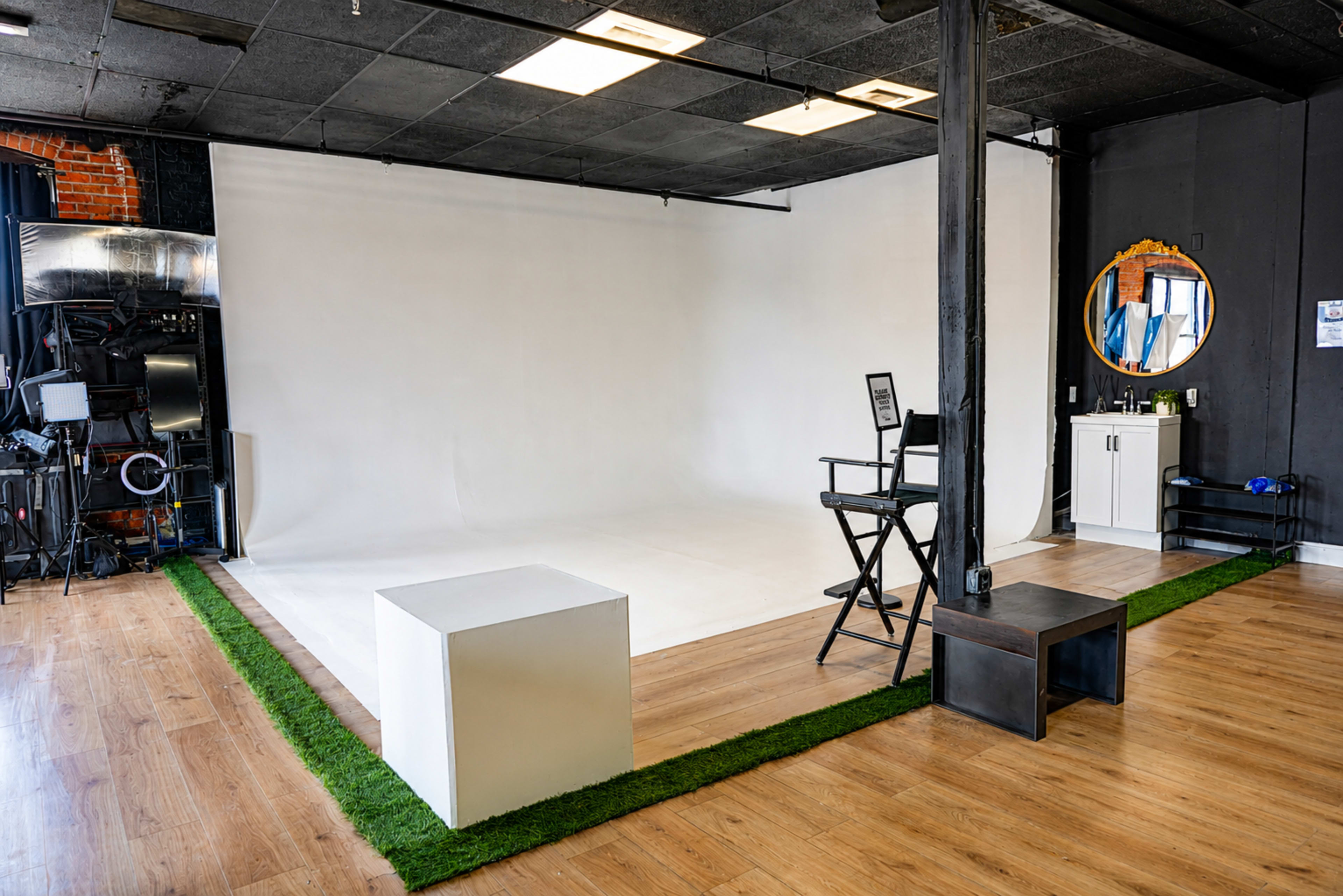The image shows a photography studio with a plain white backdrop, wooden flooring, and two pieces of furniture: a black director's chair and a small white cube on a grassy area.