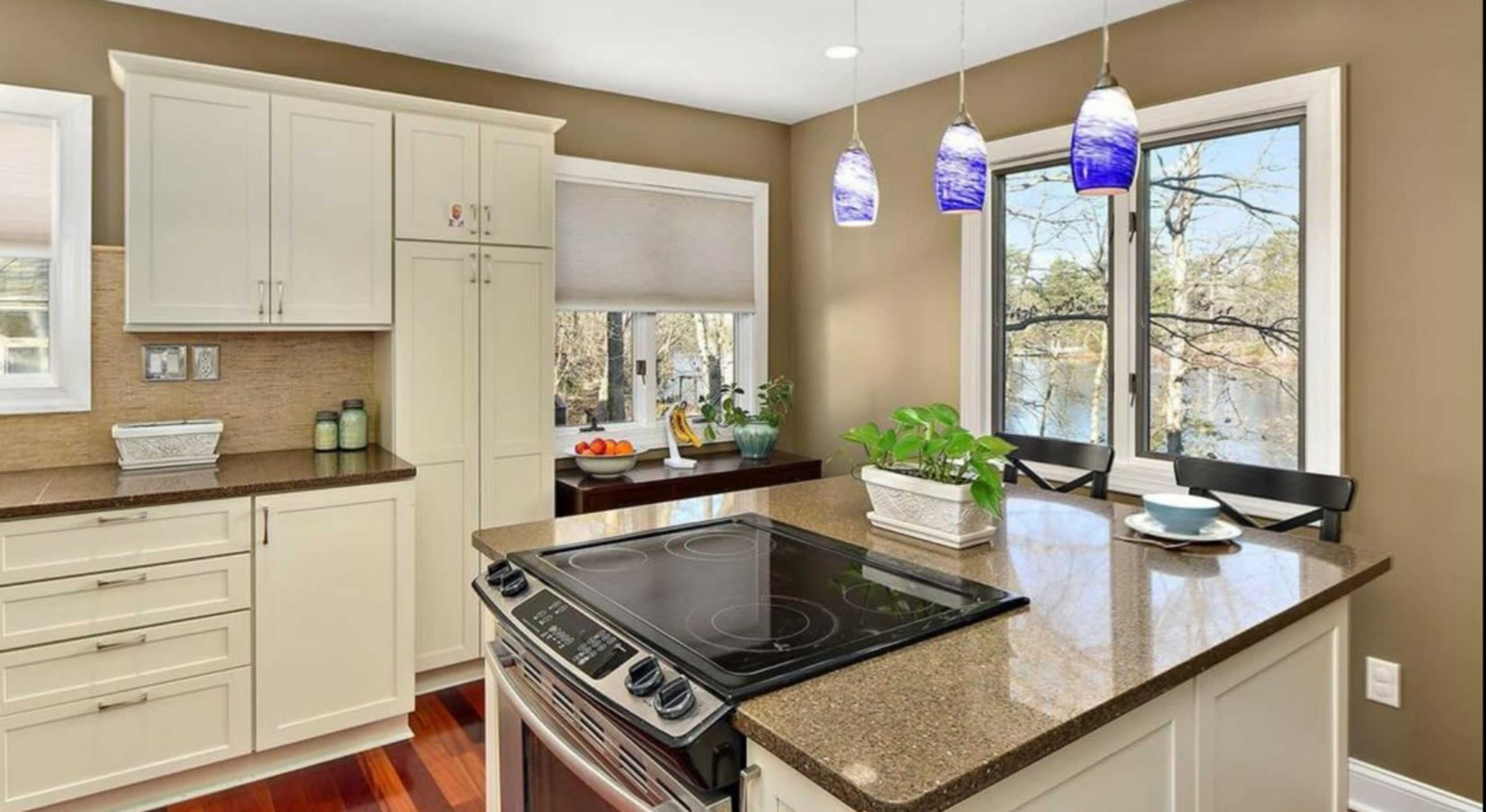 The kitchen features a central island with a stovetop, light-colored cabinetry, and windows overlooking a scenic outdoor view.