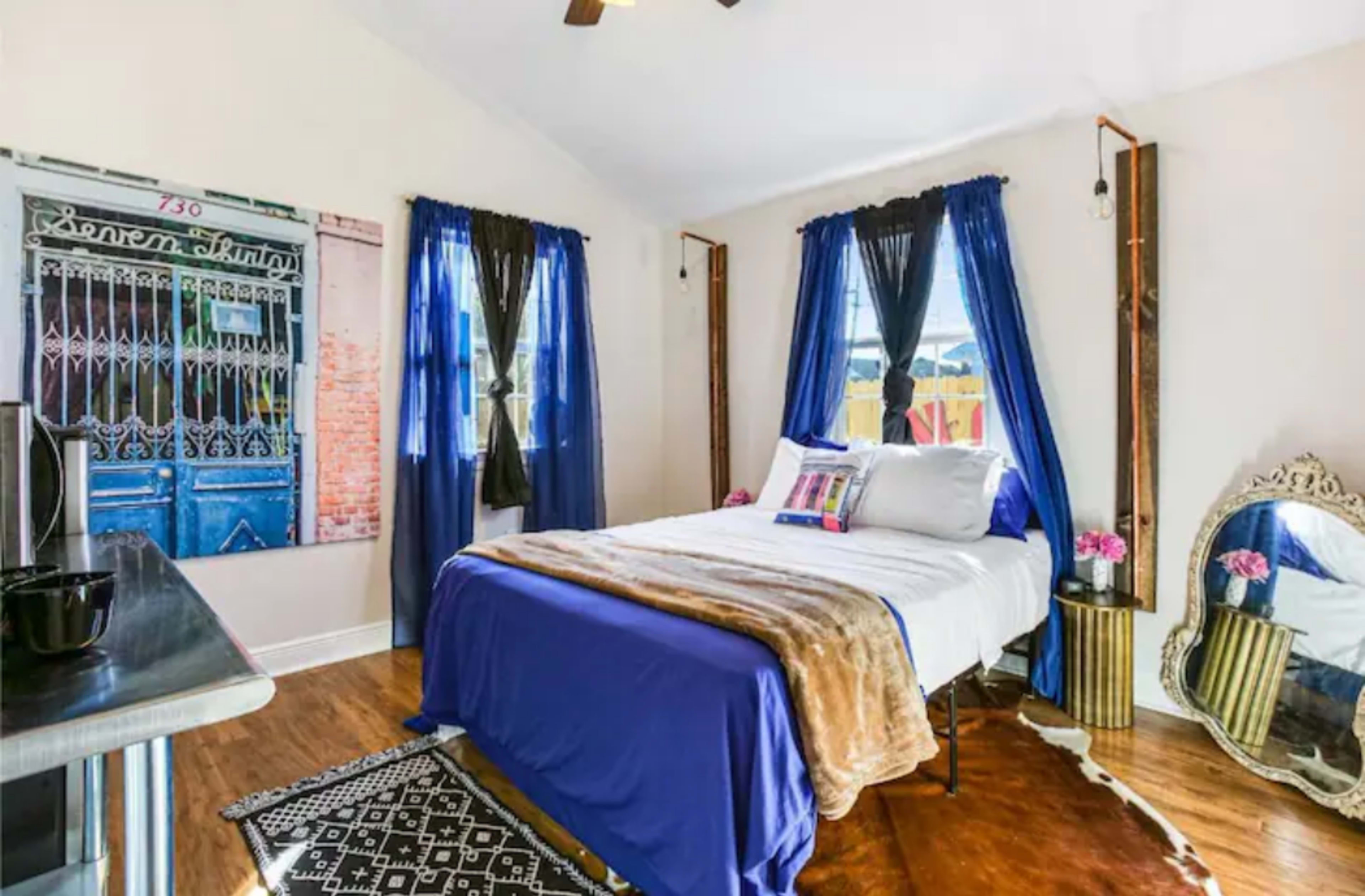 A decorated bedroom with a bed covered in a blue and brown blanket, two windows with dark blue curtains, and a vintage mirror next to a small table.