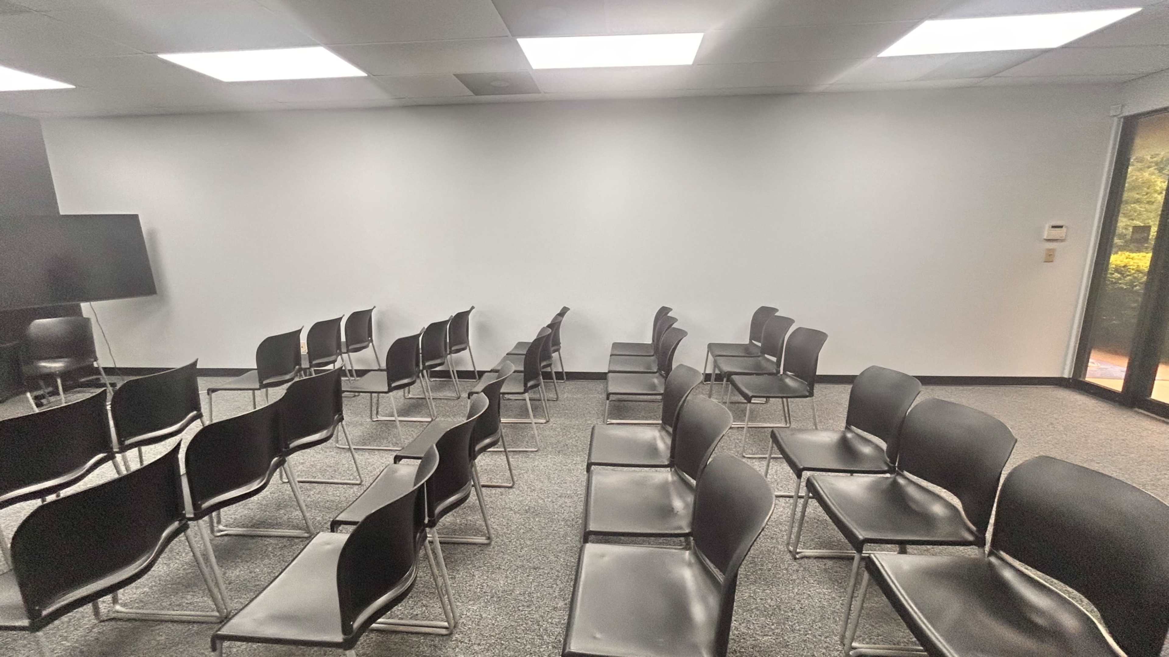 The image shows a conference room with several rows of black chairs arranged in a neat layout, facing a blank white wall.