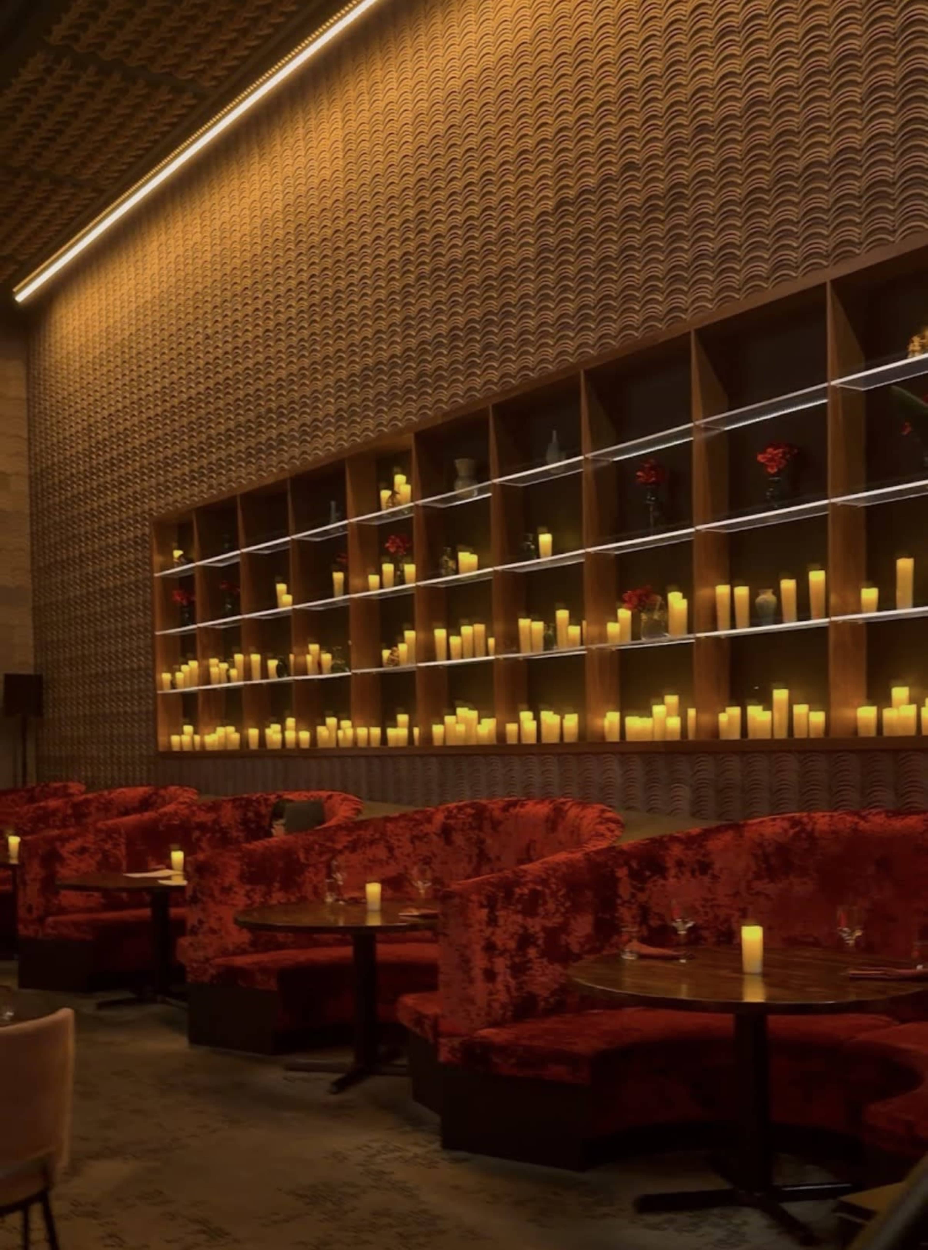 The image shows a dimly lit bar or lounge with plush red seating, decorative wooden shelves illuminated by candles, and a textured wall in the background.