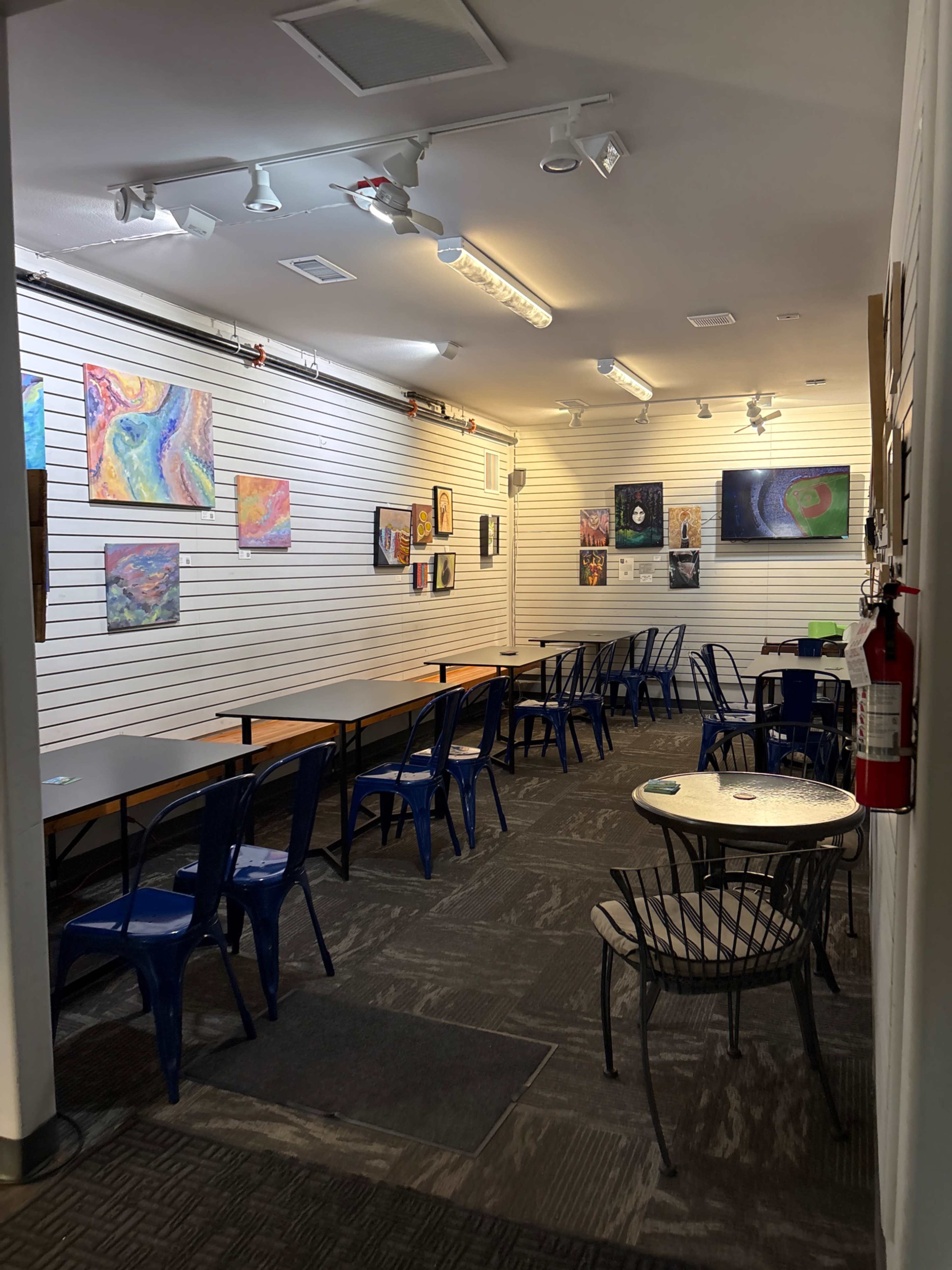 A well-lit art gallery features rows of tables and chairs, with colorful artwork displayed on the walls.