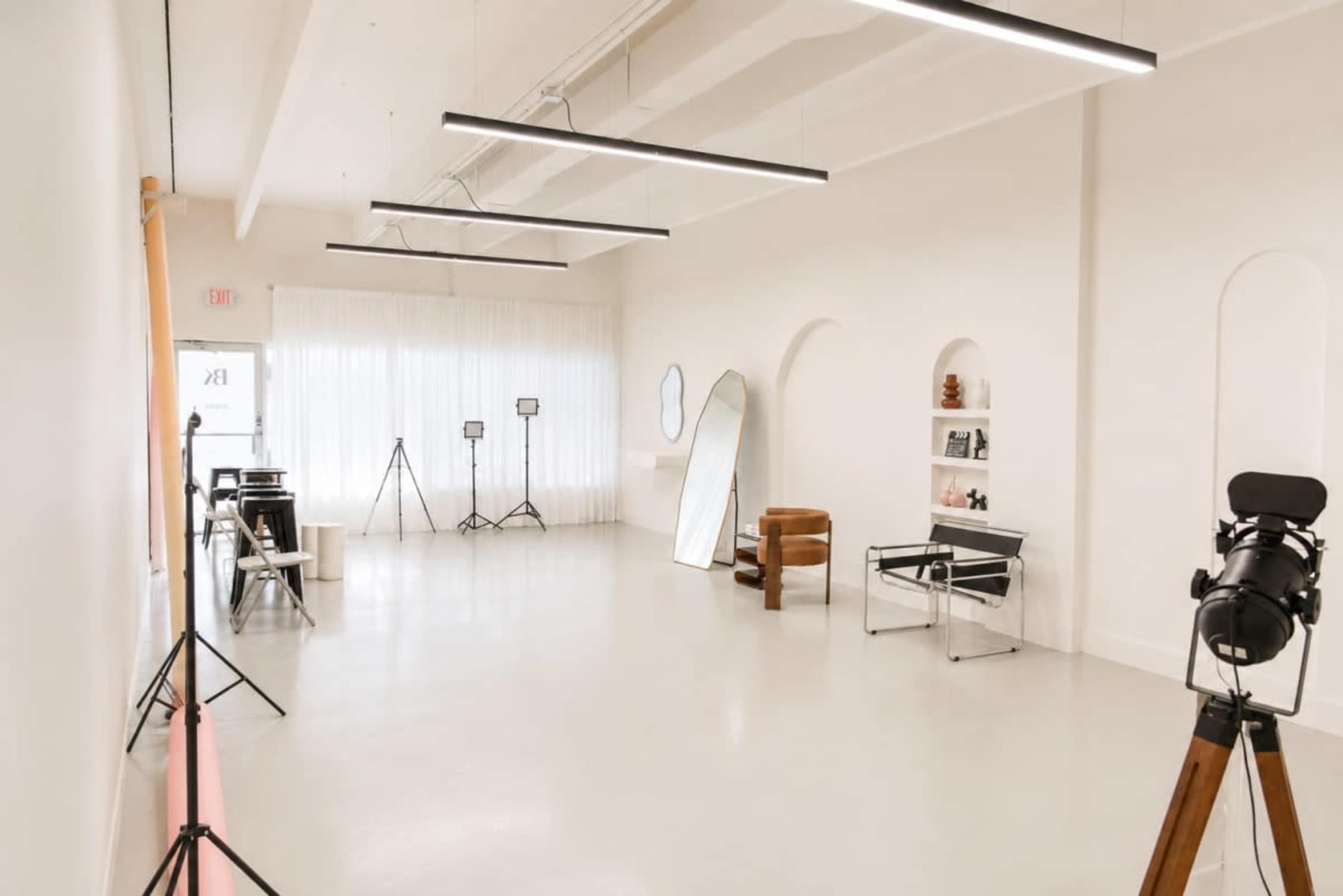 The image shows a spacious, well-lit studio featuring minimalistic furniture, photography equipment, and large windows with sheer curtains.