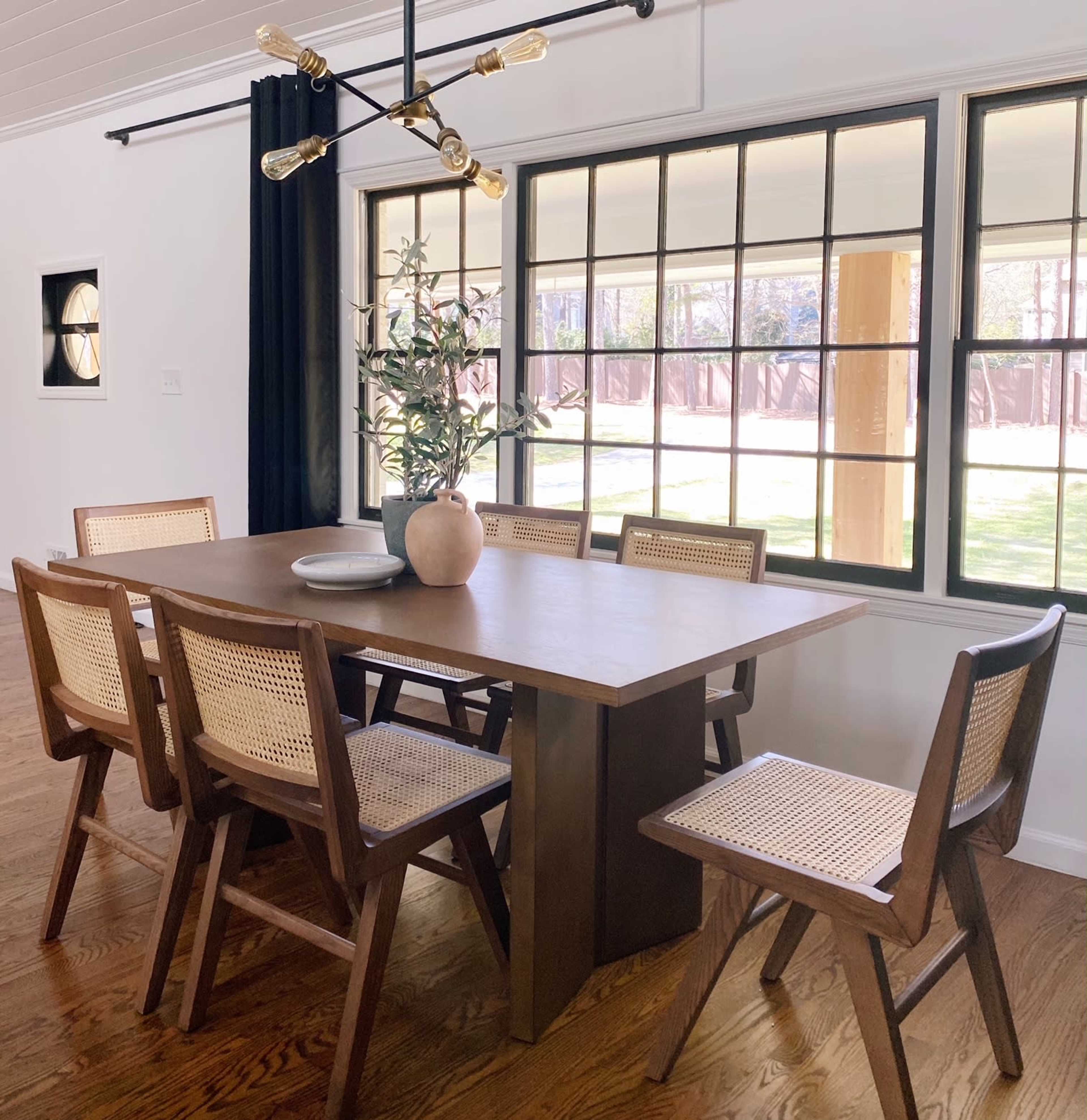 A wooden dining table with six chairs is positioned by large windows that overlook a grassy area.