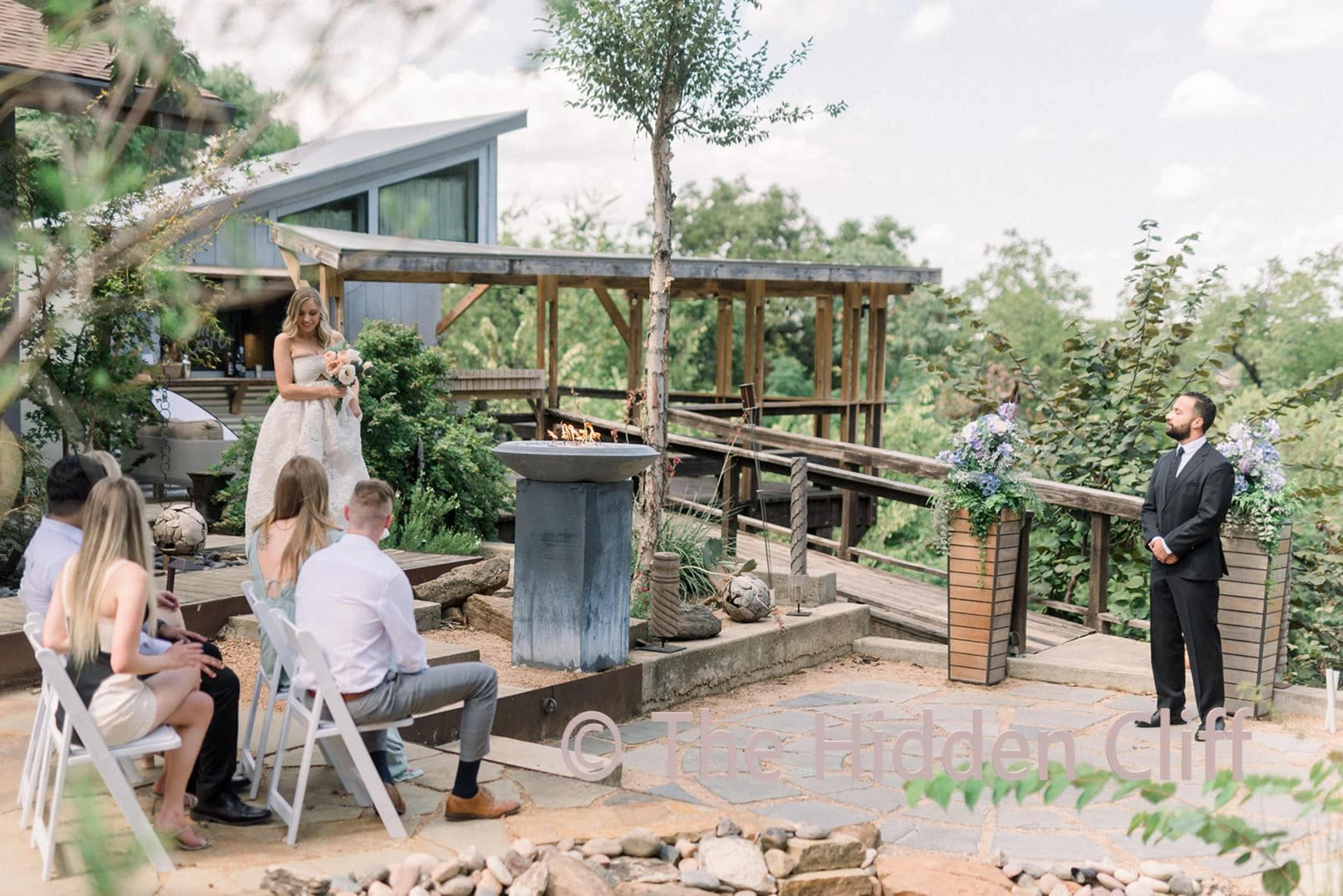 A bride stands with a bouquet in a garden ceremony while a groom faces her, surrounded by guests seated on white chairs.