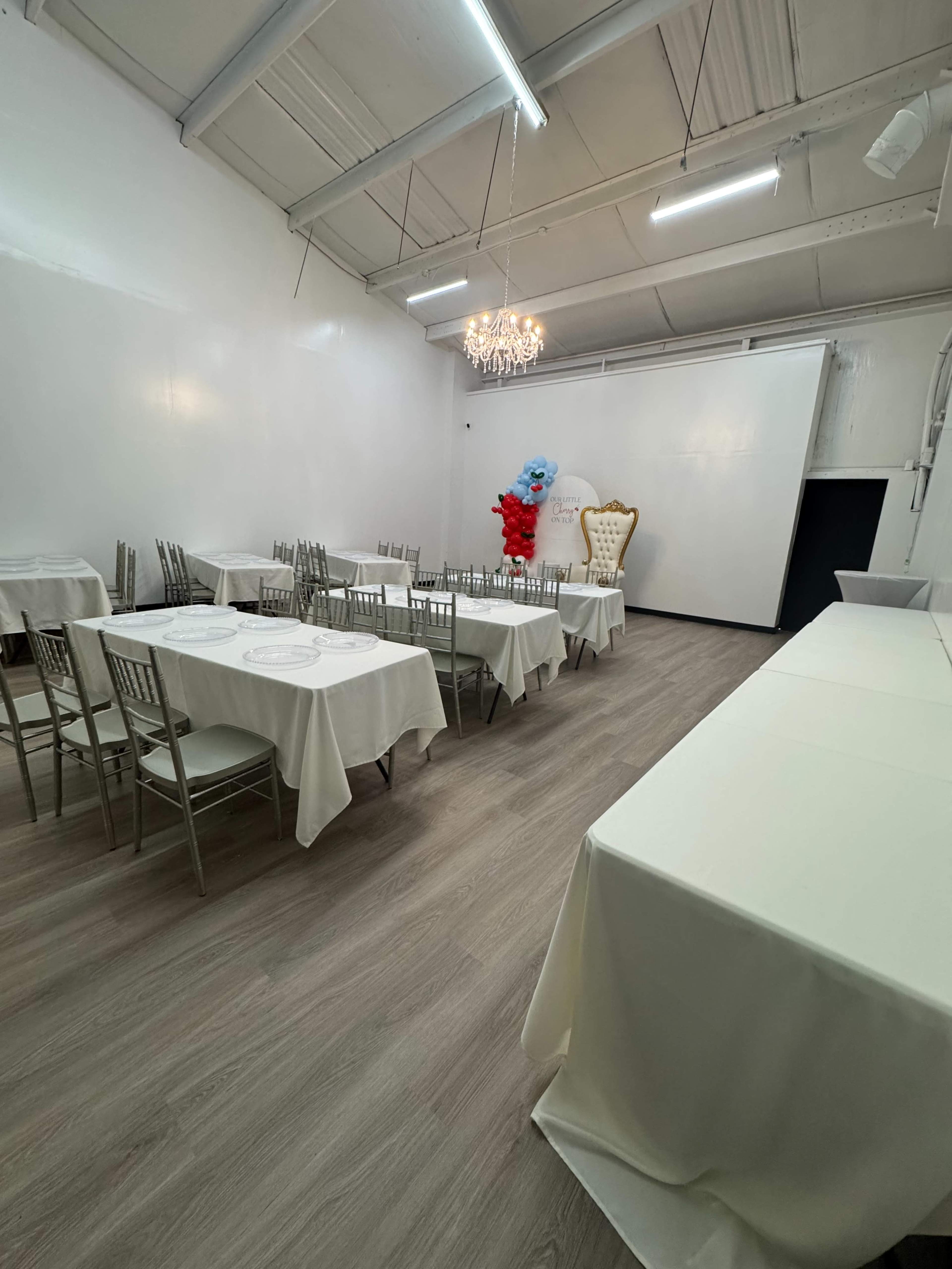 The image shows a spacious event room with white tables covered in tablecloths, arranged in rows, and decorated with a chandelier and colorful balloon arrangements at one end.