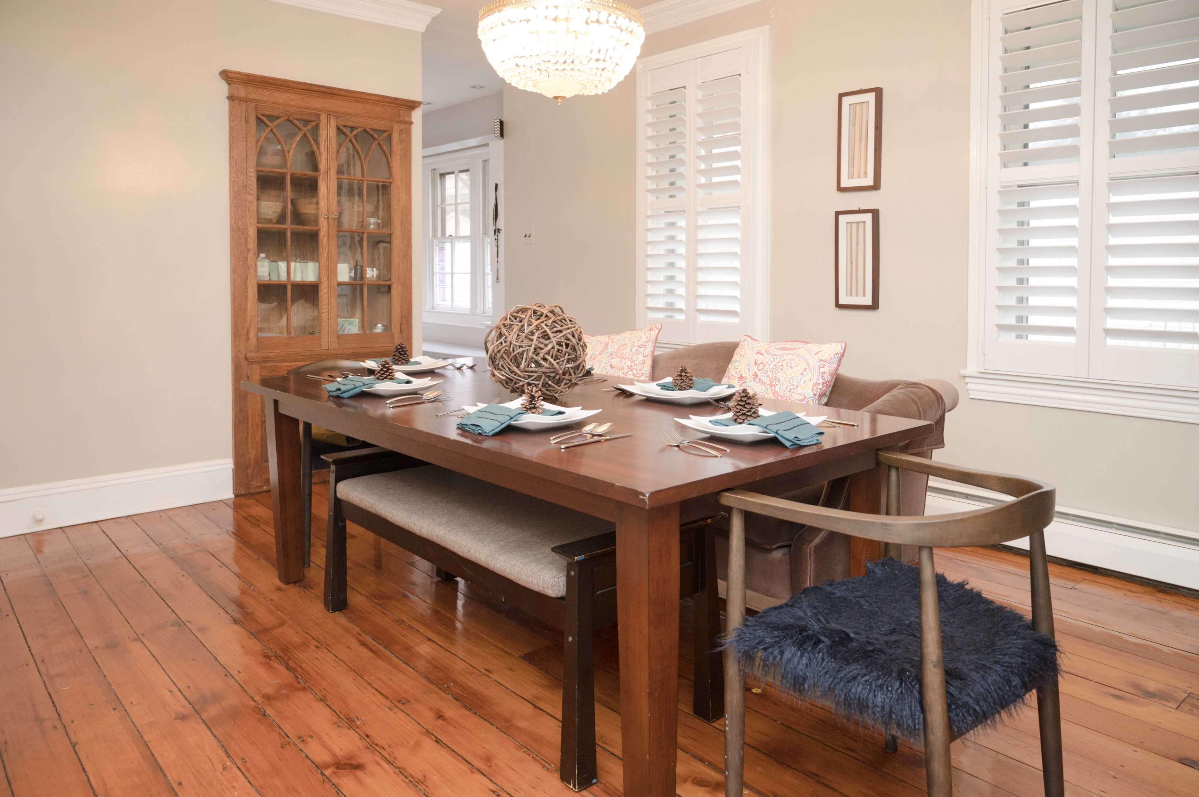 A dining room features a wooden table set for a meal, with upholstered chairs, a decorative bowl in the center, and a glass cabinet displaying items in the background.