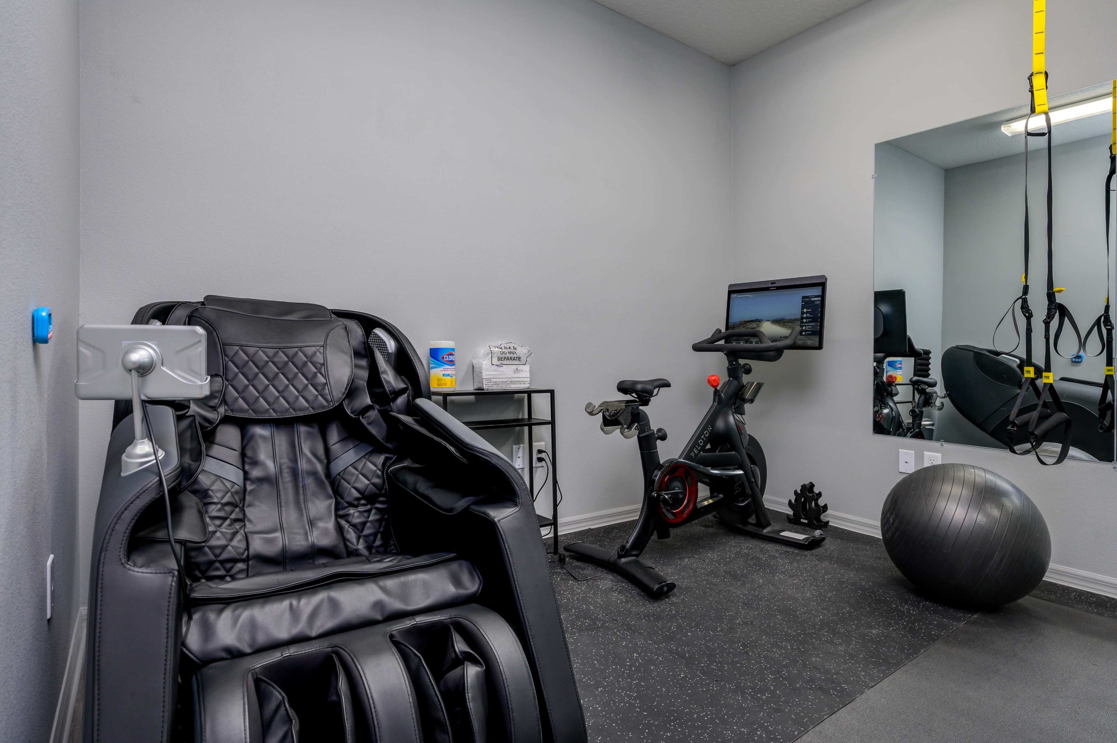 The image shows a home gym featuring a massage chair, an exercise bike, a stability ball, and workout equipment against a gray wall.