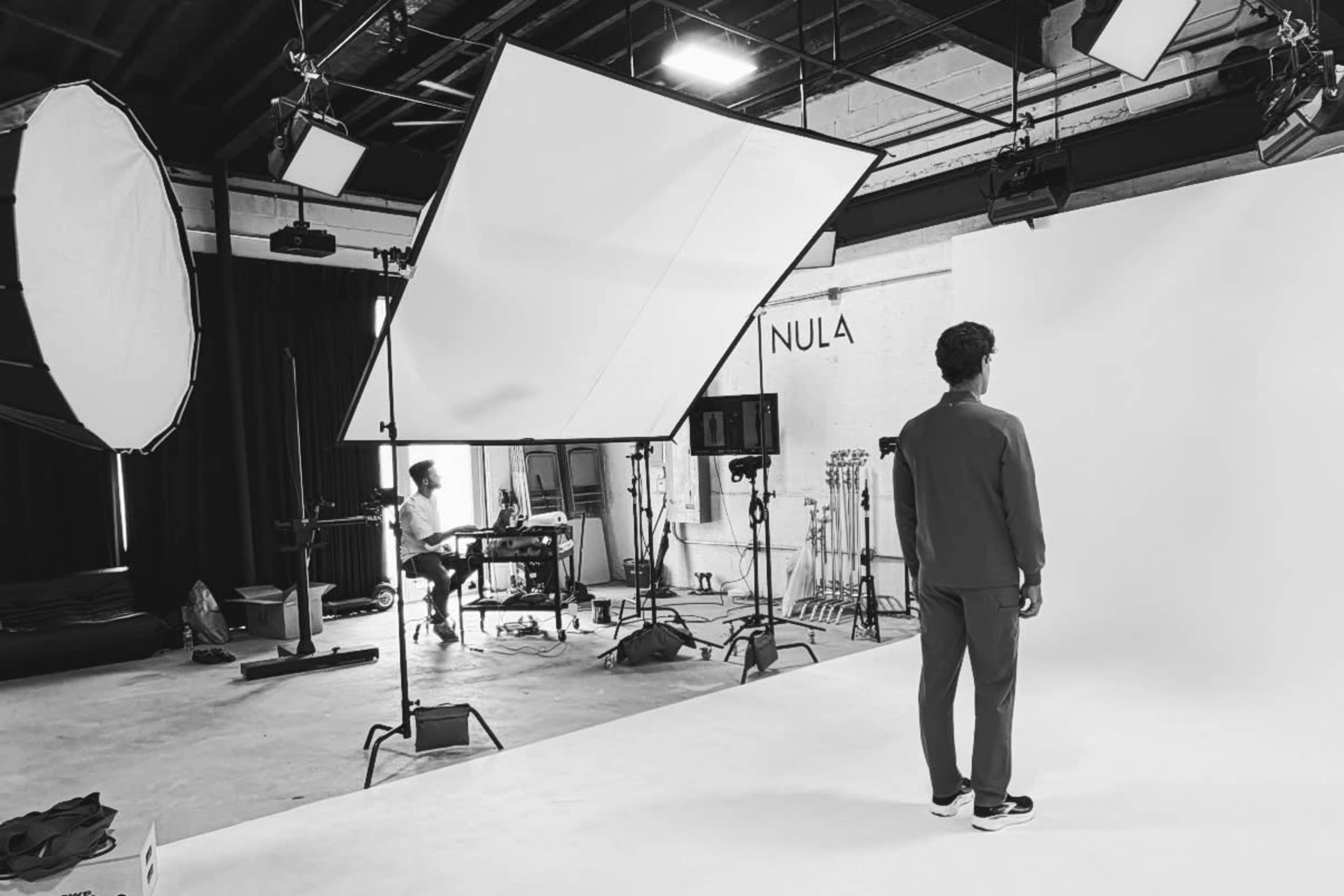 A person stands facing a blank backdrop in a well-lit photography studio with various lighting equipment and a stylist working in the background.